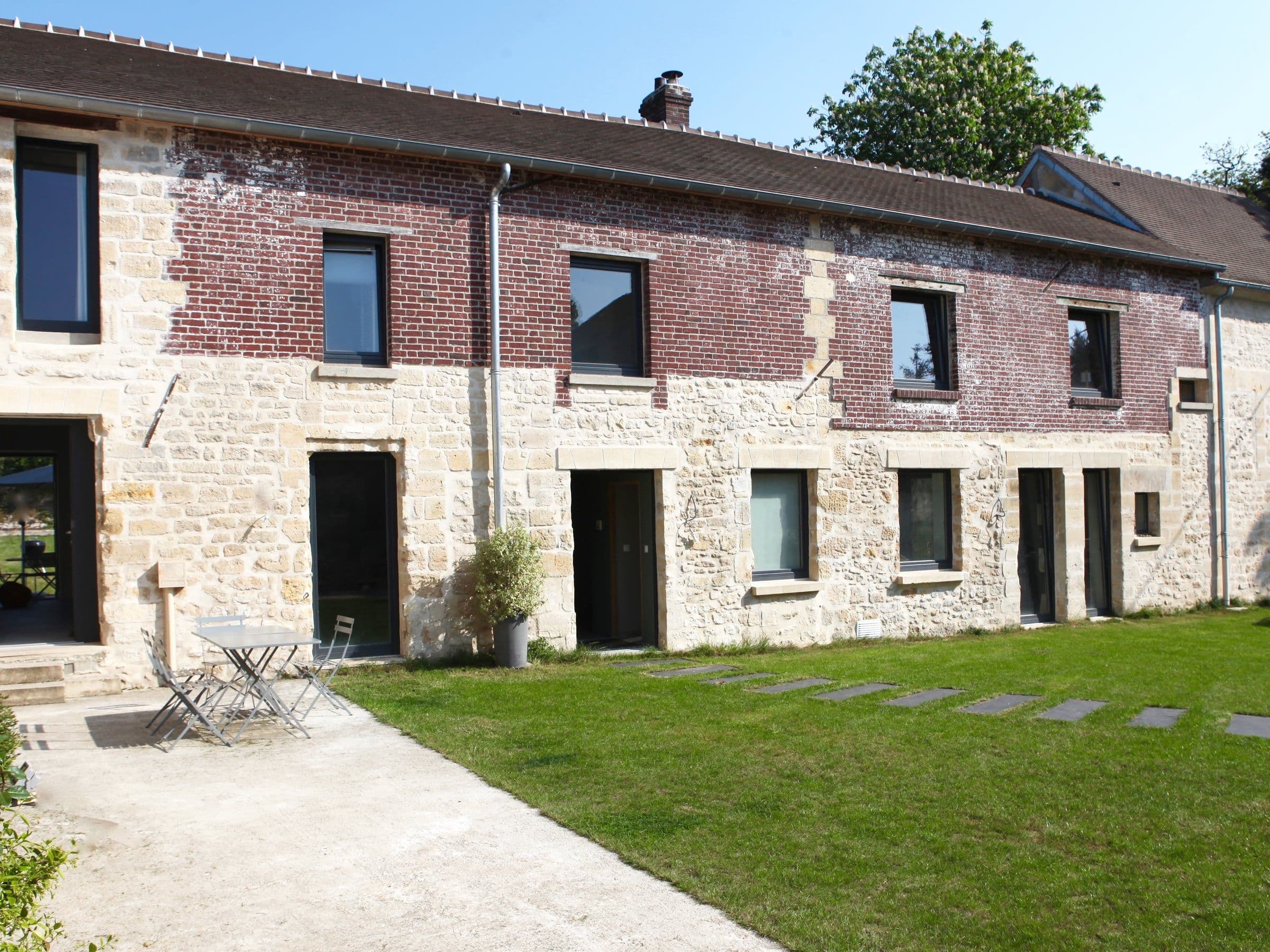 Après travaux : façade rénovée en pierre et brique rouge, équipée de nouvelles menuiseries sombres, d’une terrasse minérale et d’une pelouse.