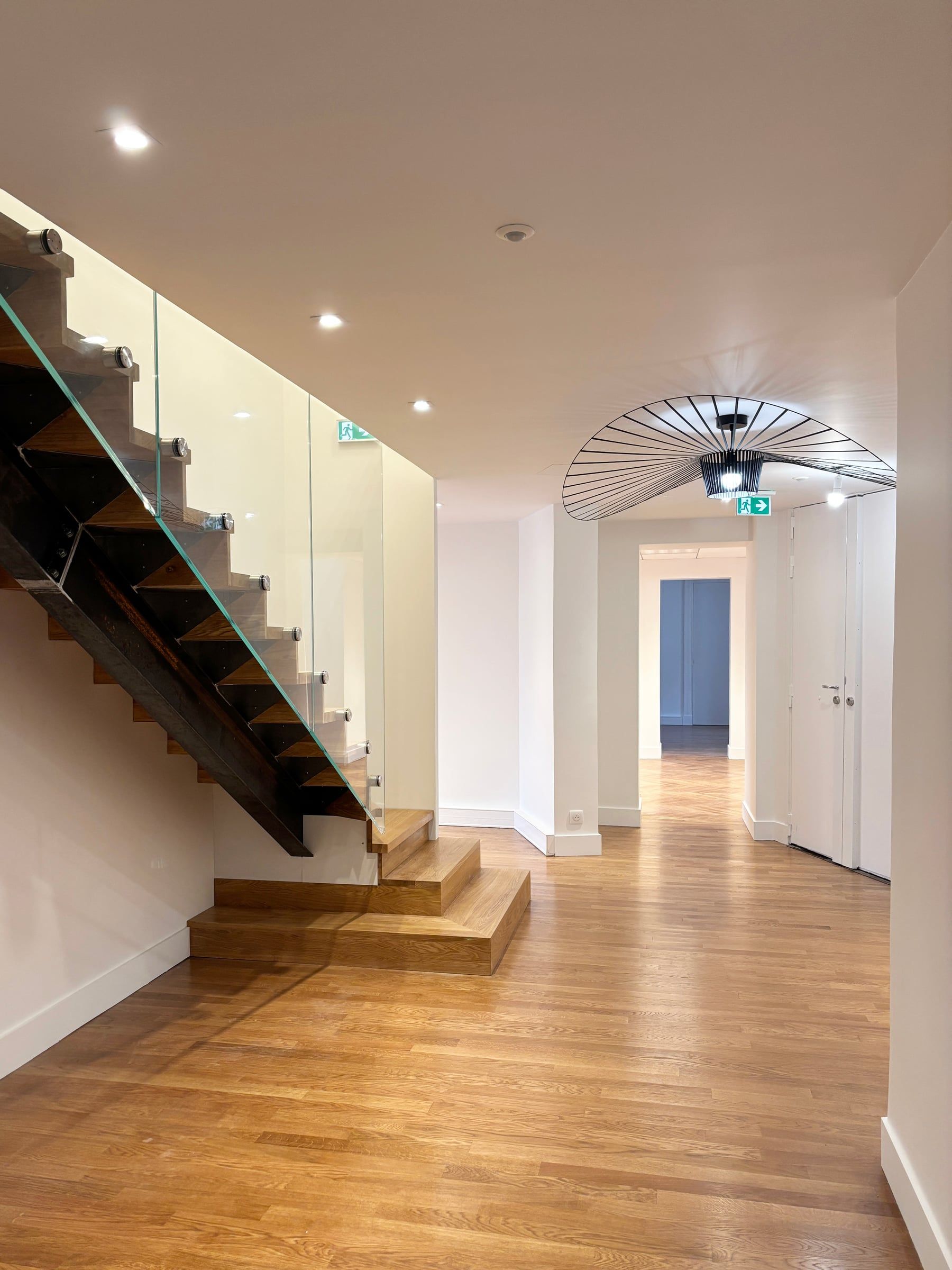 Hall de bureaux avec escalier en chêne et métal noir, garde-corps en verre, parquet clair et suspension Vertigo, aménagement de bureaux contemporain