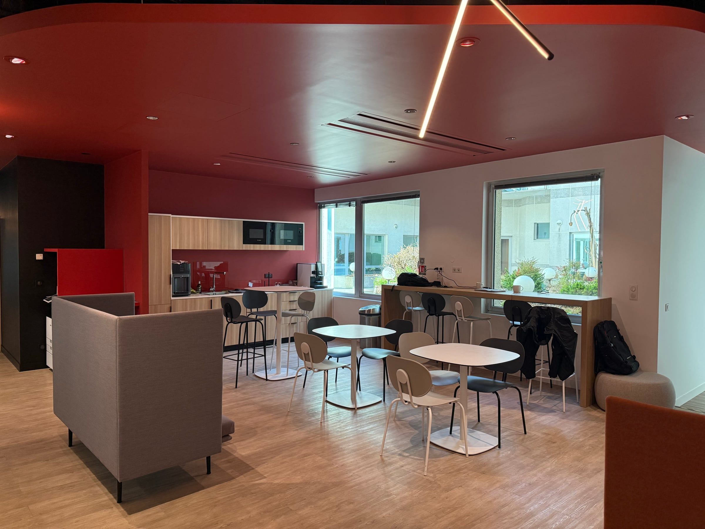 Cafétéria d'entreprise au plafond et mur rouge terracotta, coin cuisine, tables rondes et chaises design sur sol en parquet clair.