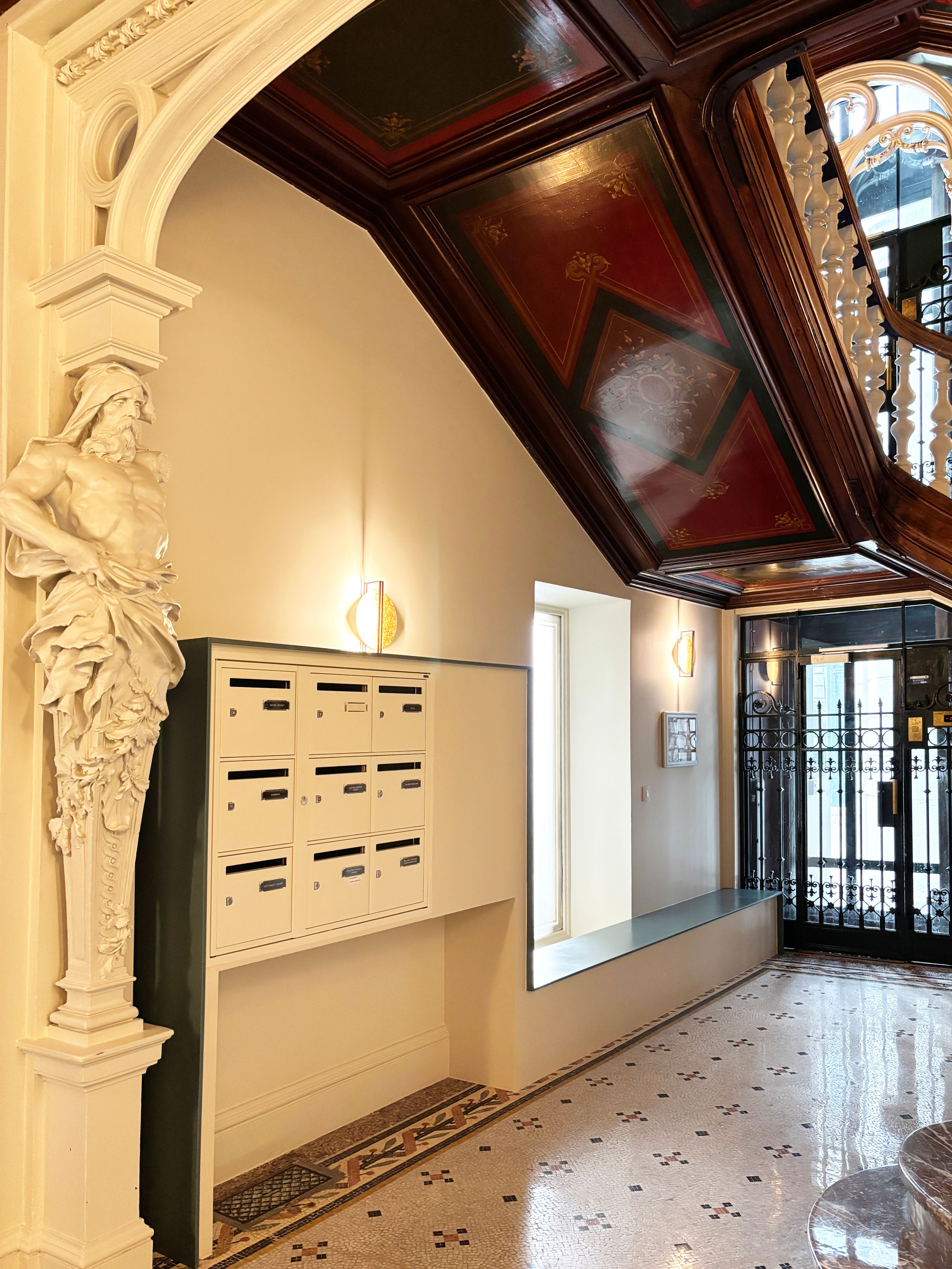 Entrée d’immeuble sous l’escalier, plafond peint restauré, boîtes aux lettres encastrées et porte en fer forgé sur sol mosaïque.