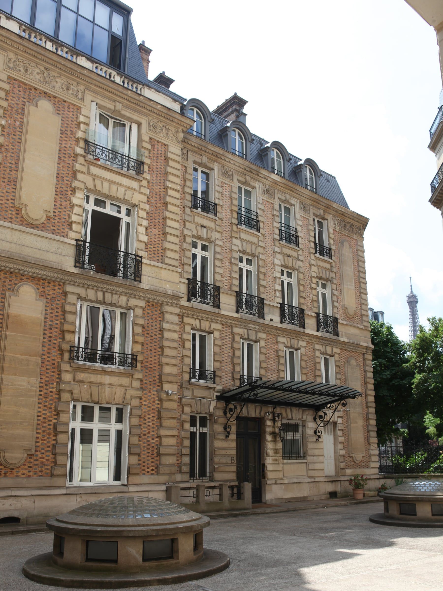 Façade haussmannienne en pierre de taille et brique sur cour pavée, marquise en fer forgé et tour Eiffel à l'arrière-plan, rénovation de maison.