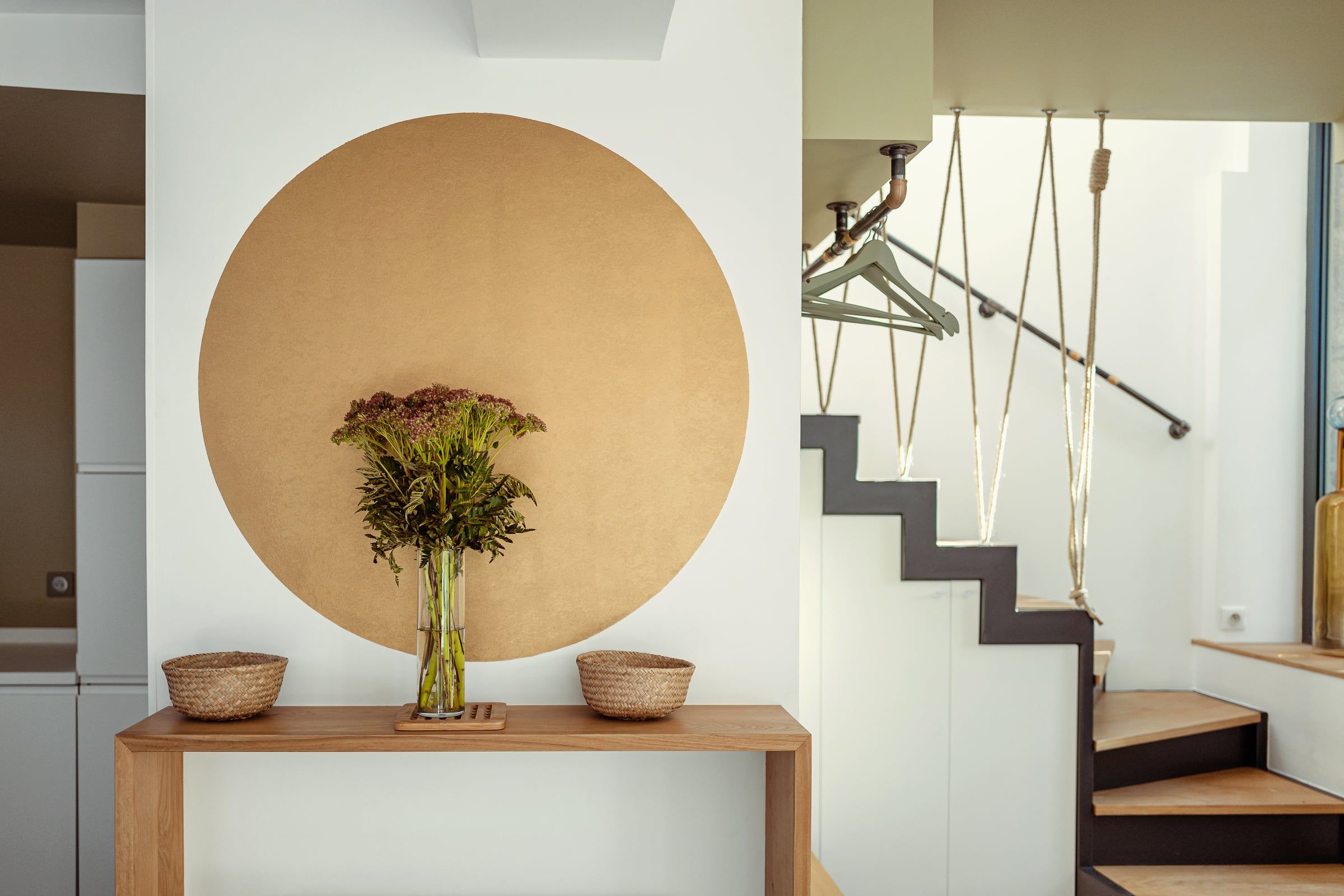 Entrée avec console en chêne devant un grand disque ocre peint et escalier contemporain suspendu par des cordages en chanvre, architecture intérieure soignée.