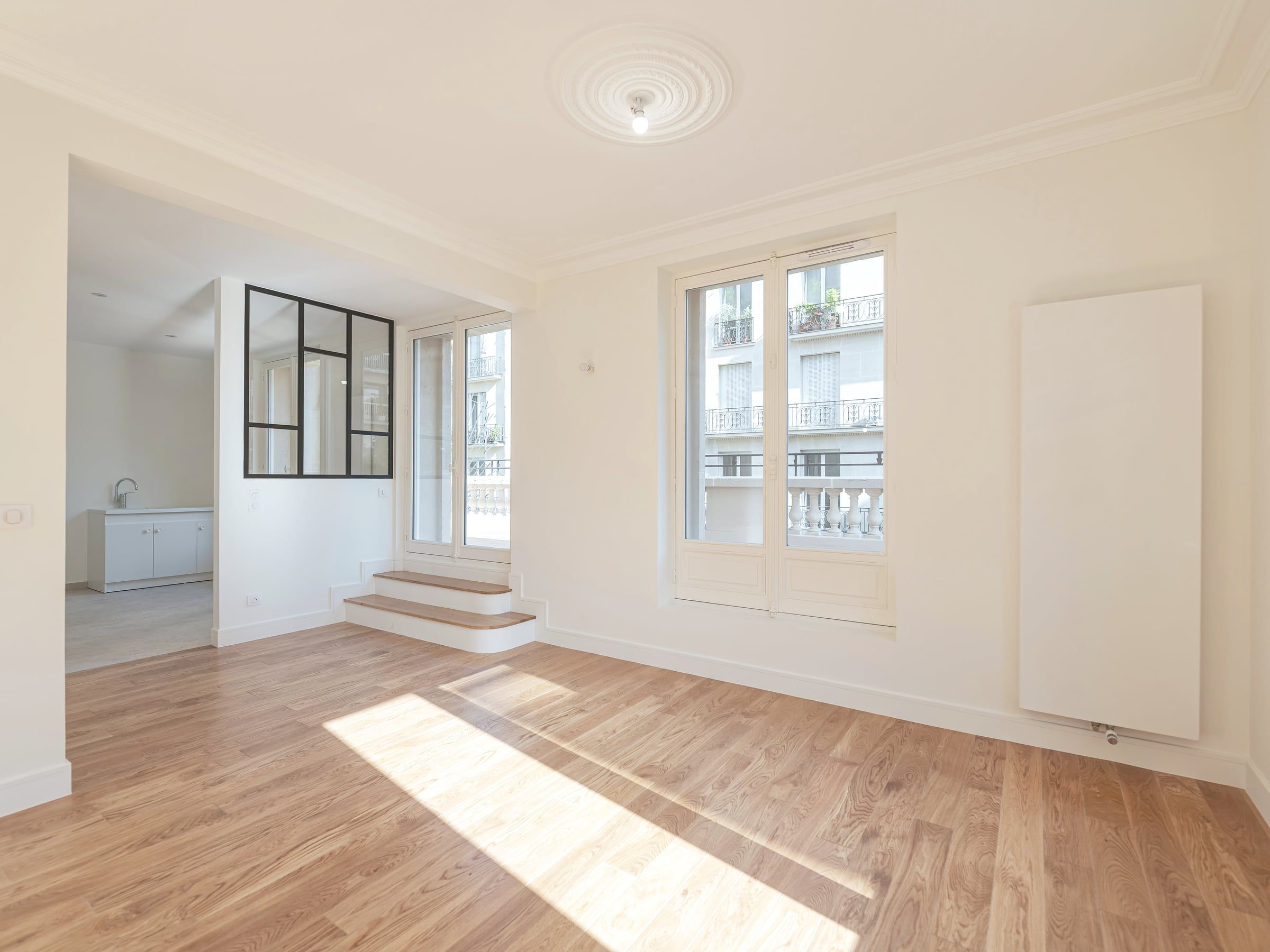 Séjour résidentiel rénové par un architecte d’intérieur, avec parquet en chêne, rosace au plafond, verrière noire et portes-fenêtres sur balcon.