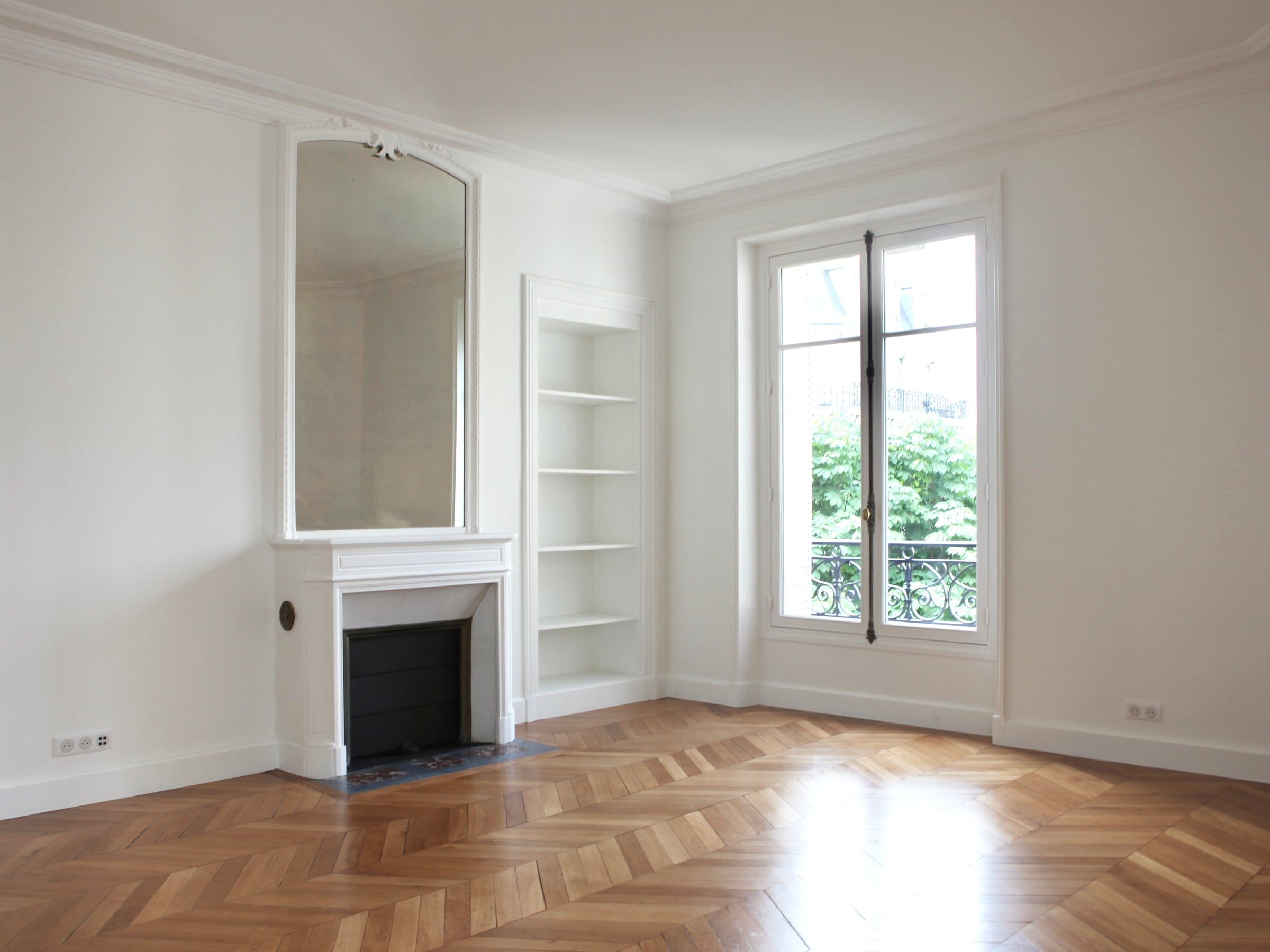 Pièce haussmannienne aux murs blancs après rénovation d'appartement, cheminée en pierre à trumeau, niche d'étagères et parquet à chevrons.