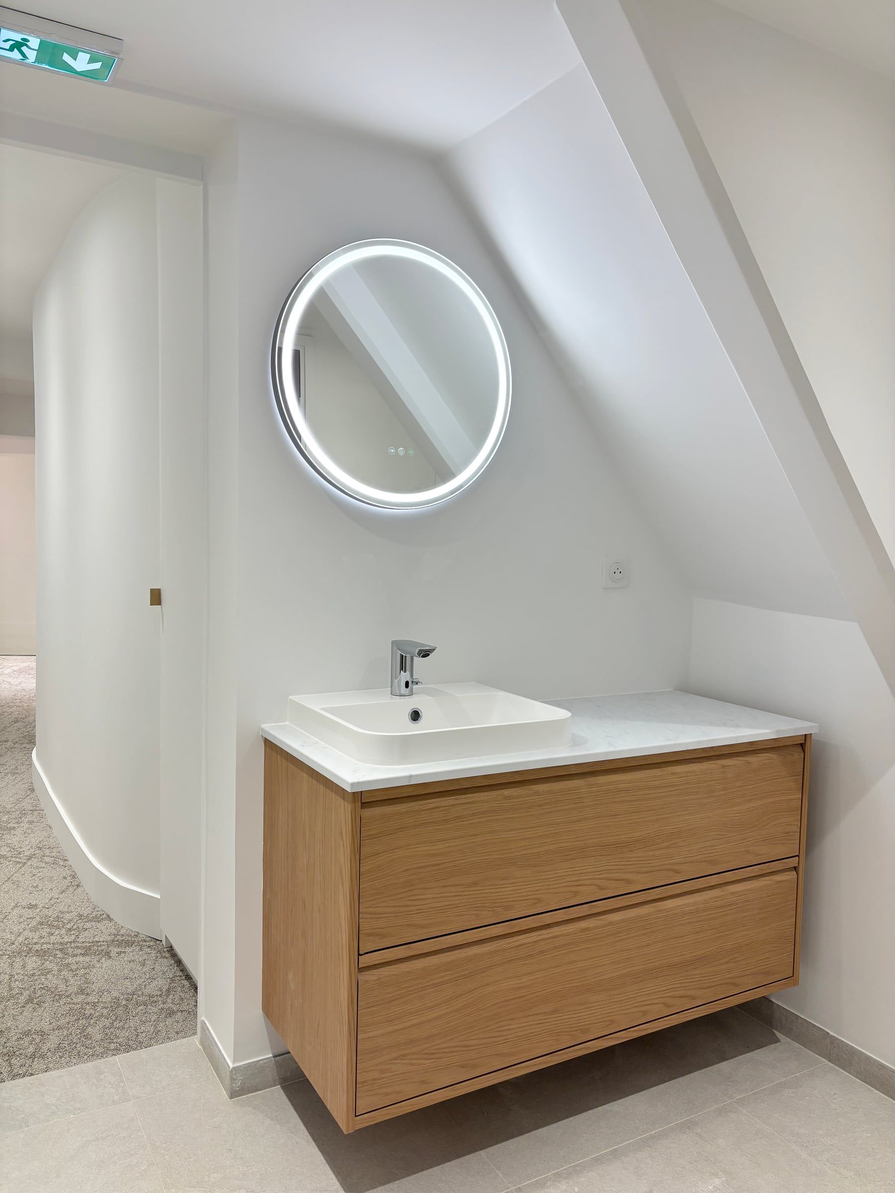 Point d'eau de bureaux sous comble avec vasque carrée sur meuble suspendu en chêne et miroir rond rétroéclairé, aménagement de bureaux soigné