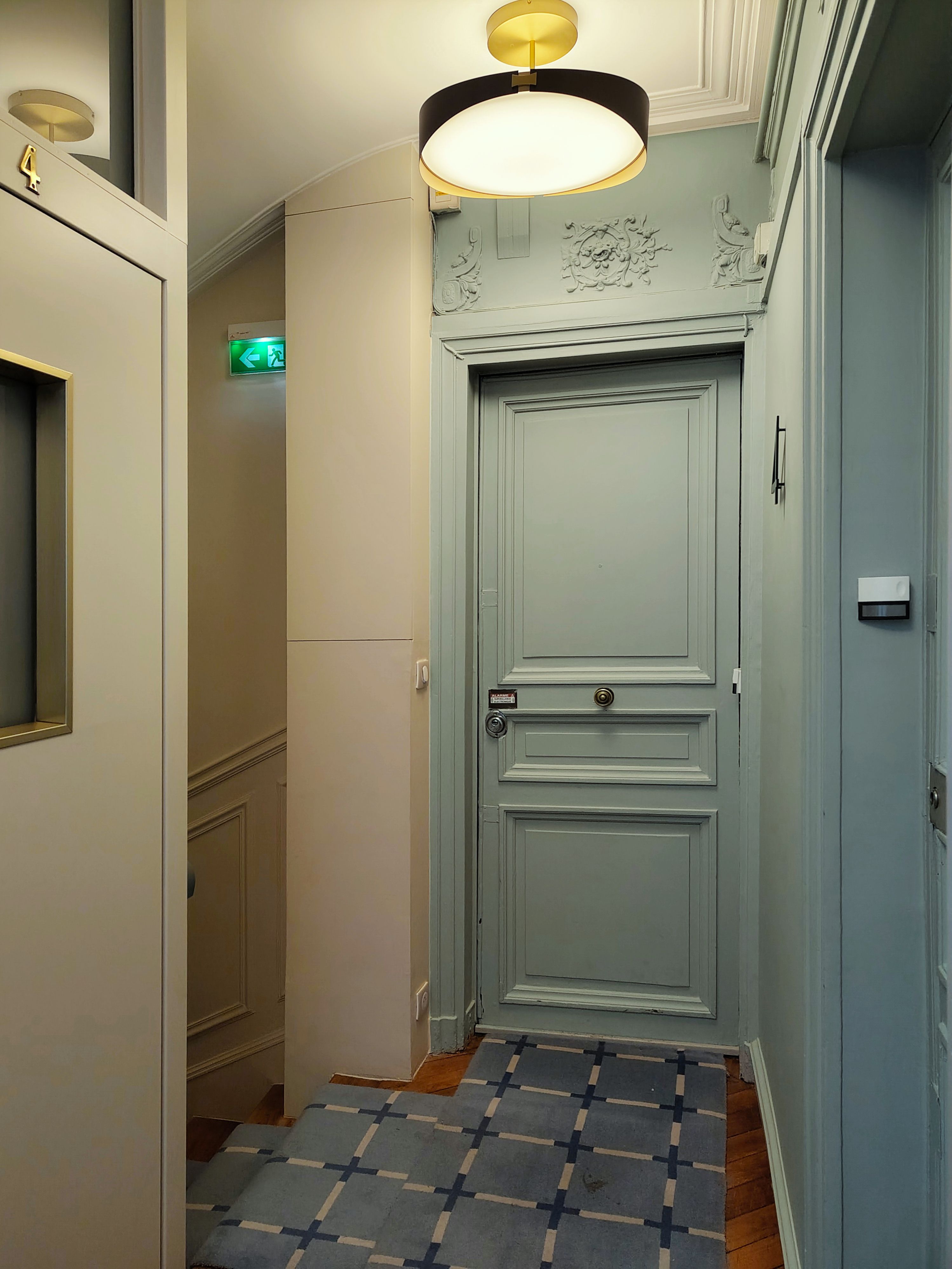 Cage d'escalier hélicoïdale aux murs à panneaux moulurés, marches en chêne habillées d'un tapis bleu à motif de croix et corniche en plâtre.