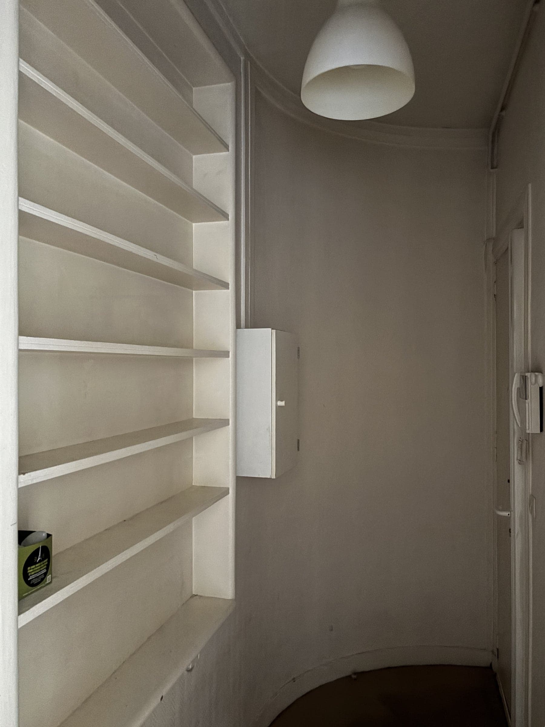 Avant travaux : débarras incurvé aux étagères murales en bois peint, mur arrondi et suspension opaline dans un appartement ancien.