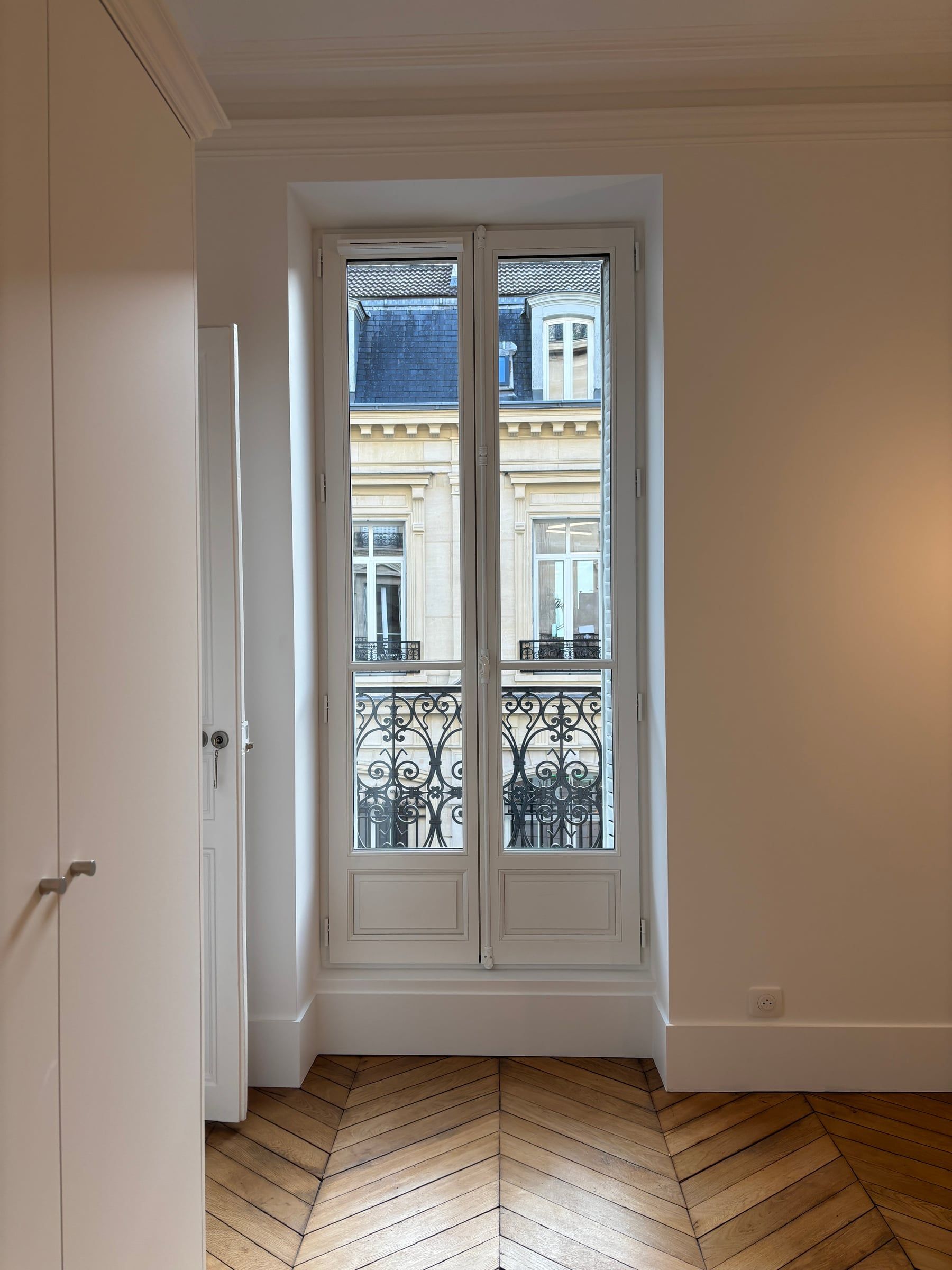 Fenêtre haussmannienne à garde-corps en fer forgé encadrée de moulures, donnant sur une façade en pierre, parquet point de Hongrie.
