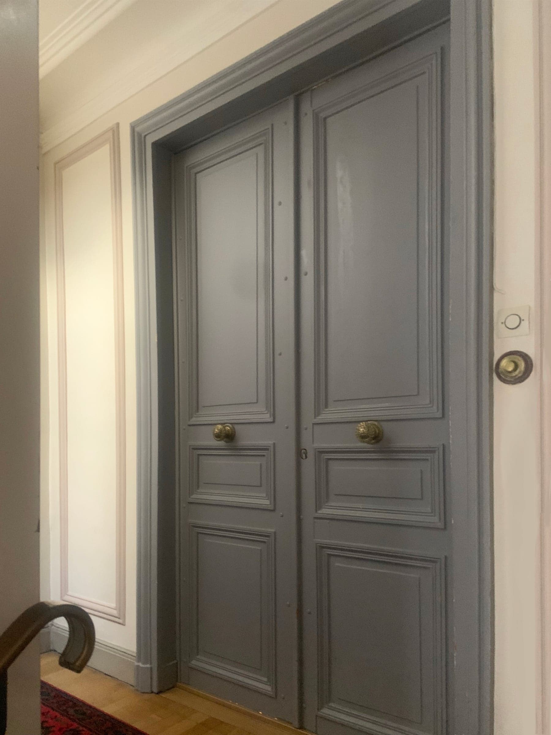 Avant travaux : palier haussmannien aux boiseries grises ternes, double porte palière à panneaux moulurés et poignées en laiton, mur à moulures défraîchies.