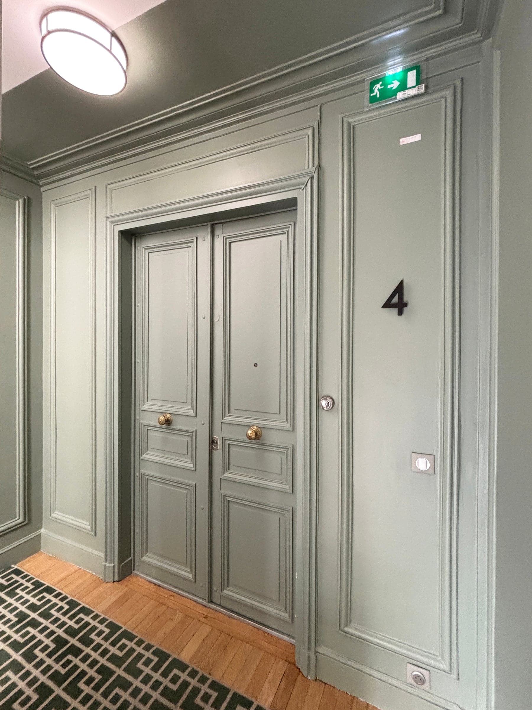 Après travaux : palier haussmannien aux boiseries vert céladon et moulures, double porte à poignées en laiton et tapis géométrique, rénovation de parties communes.