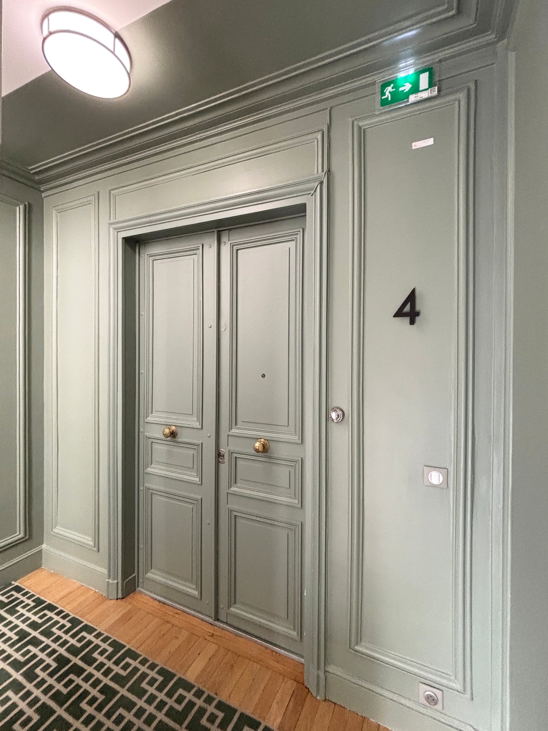 Palier haussmannien aux boiseries et moulures vert sauge, double porte palière à poignées en laiton, parquet et tapis géométrique vert, après rénovation de parties communes.