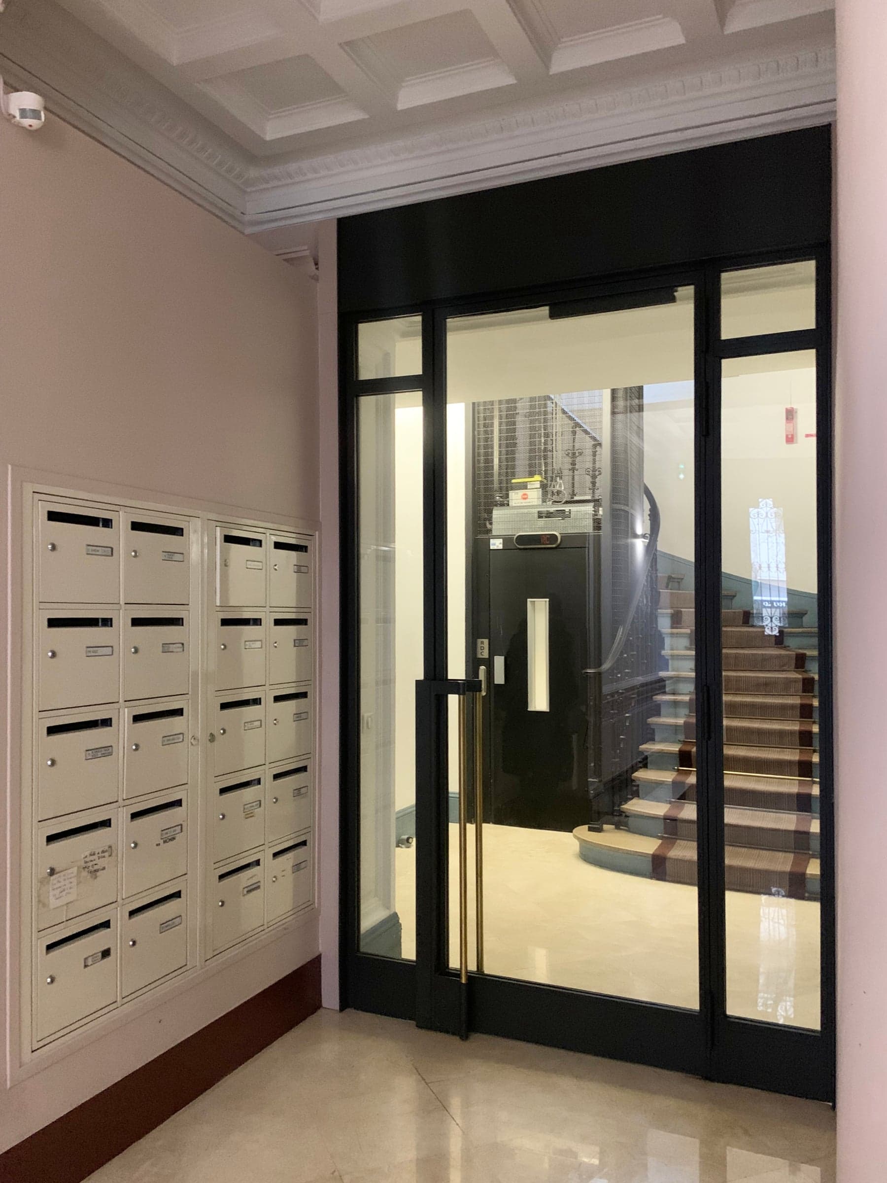 Avant travaux : hall d’immeuble aux murs roses, boîtes aux lettres beiges et porte vitrée noire donnant sur l’escalier.