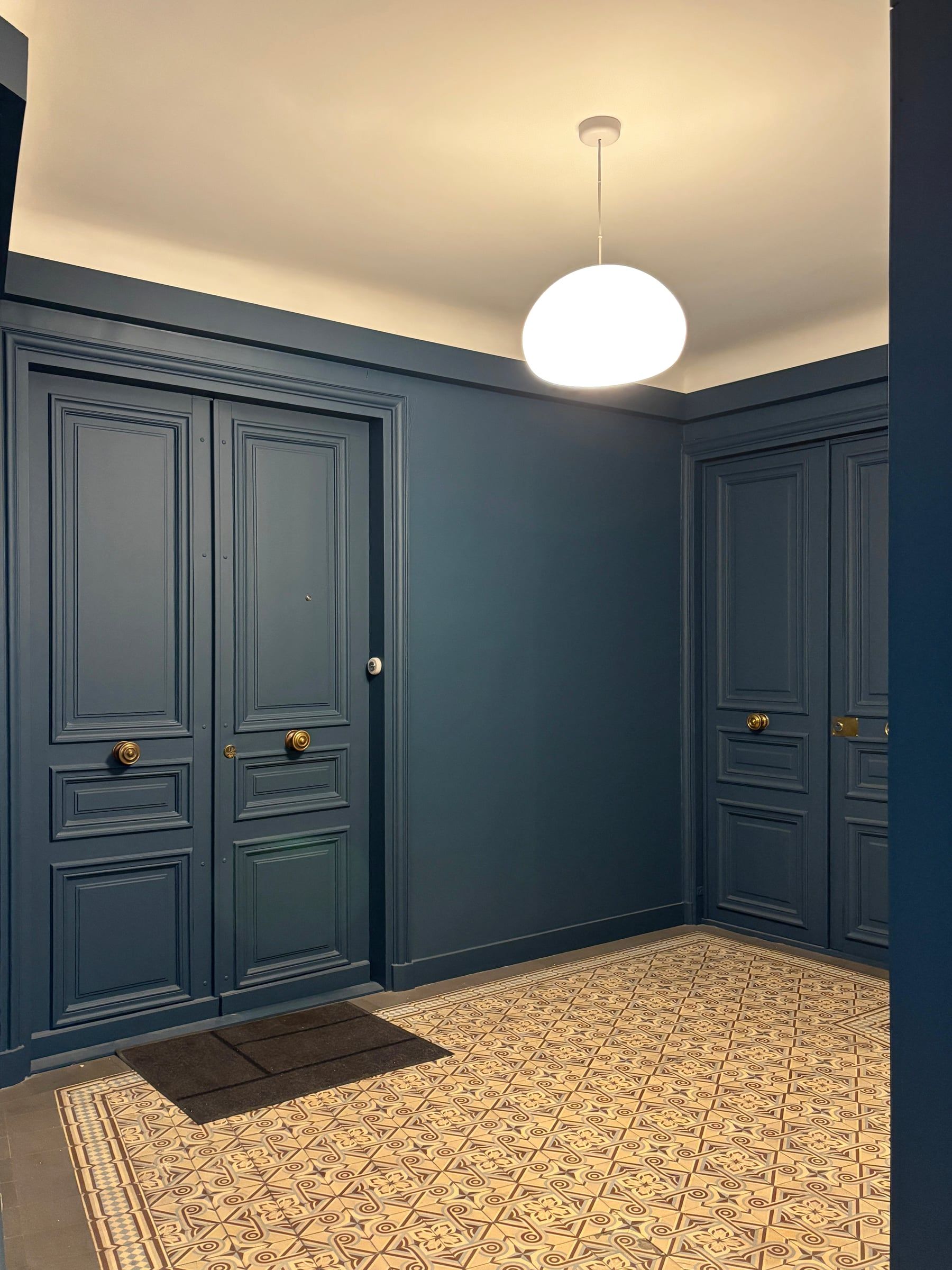 Aménagement d’entrée de copropriété avec boiseries bleu nuit, portes à panneaux moulurés et sol ancien à carreaux décorés.