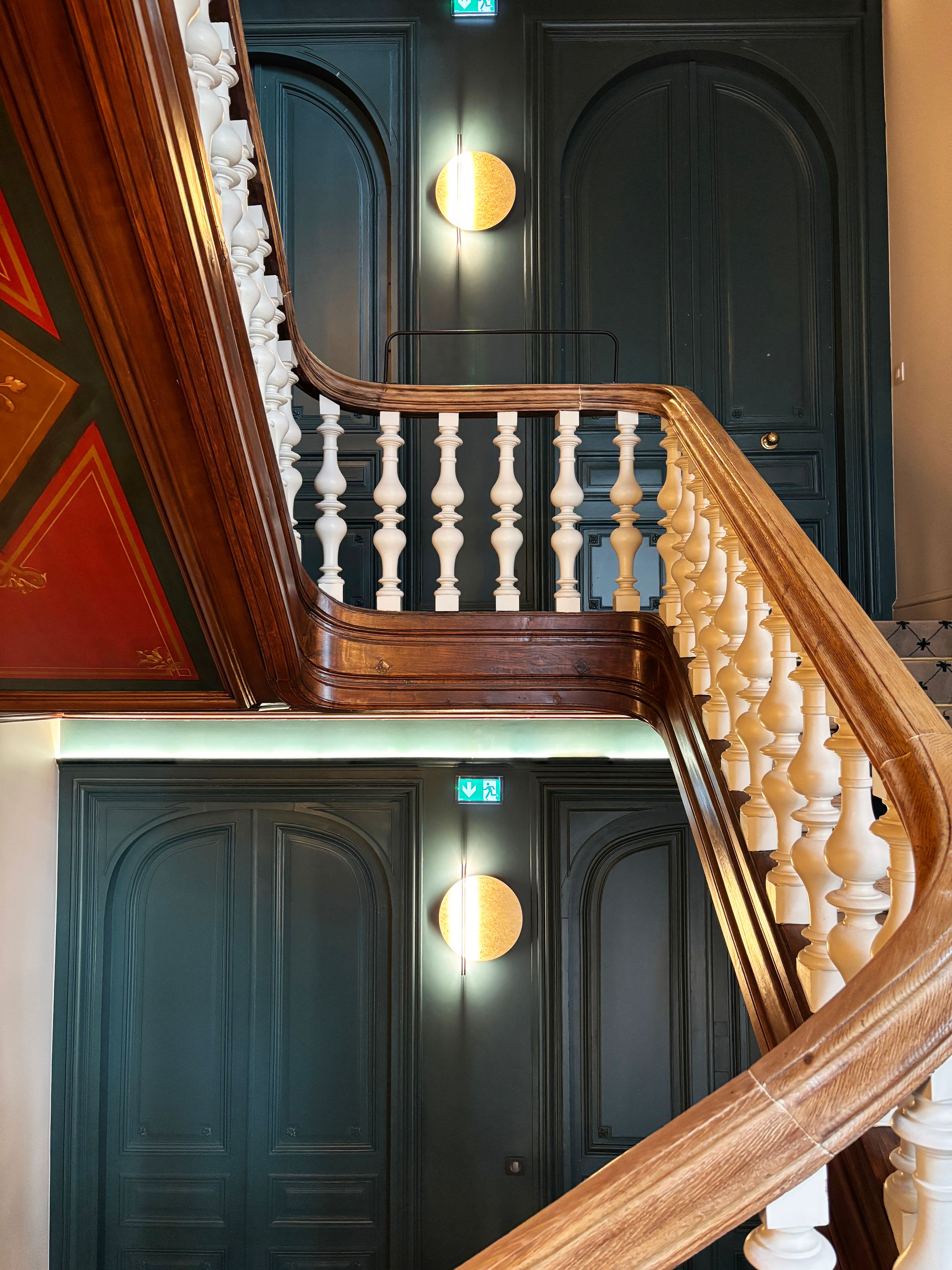 Palier de cage d’escalier aux portes moulurées vert sombre, rampe en bois et balustres blancs, avec garde-corps remis aux normes.