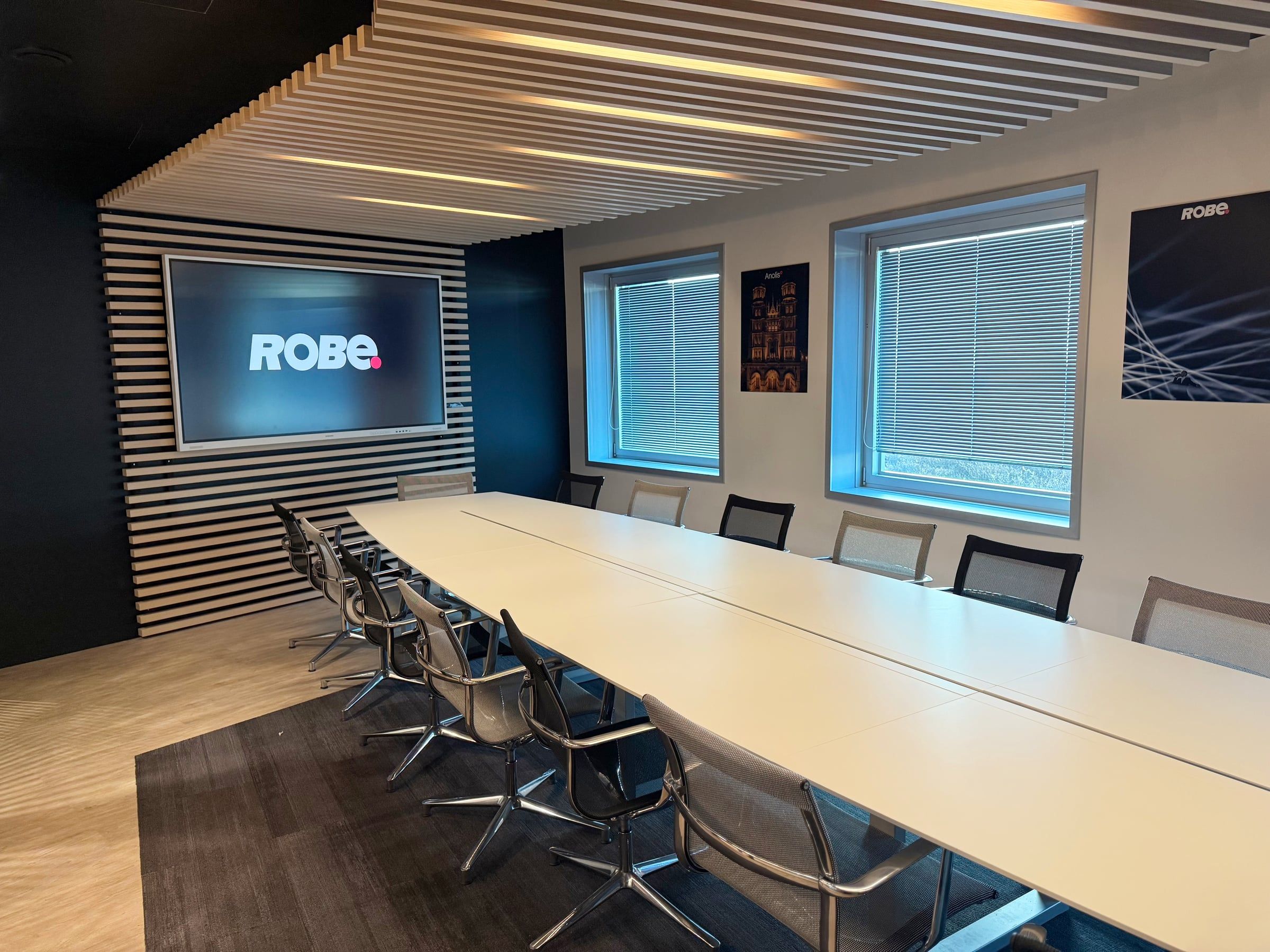 Salle de réunion aux bureaux, longue table blanche, mur d'écran encadré de tasseaux bois et faux plafond en lames acoustiques rétroéclairées.