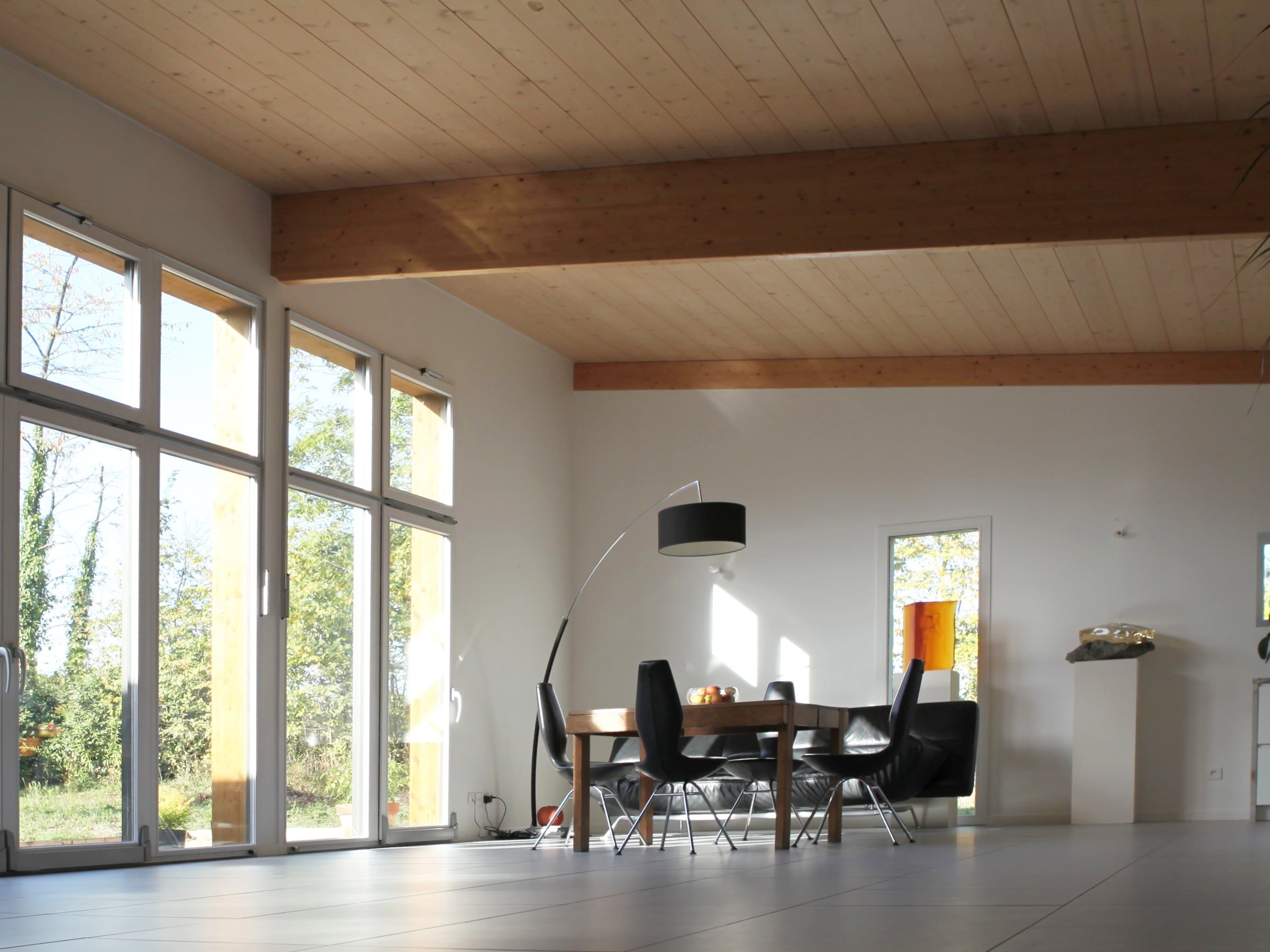 Aménagement de salle à manger dans une pièce à vivre aux grandes baies vitrées, poutres apparentes en bois et carrelage gris clair.