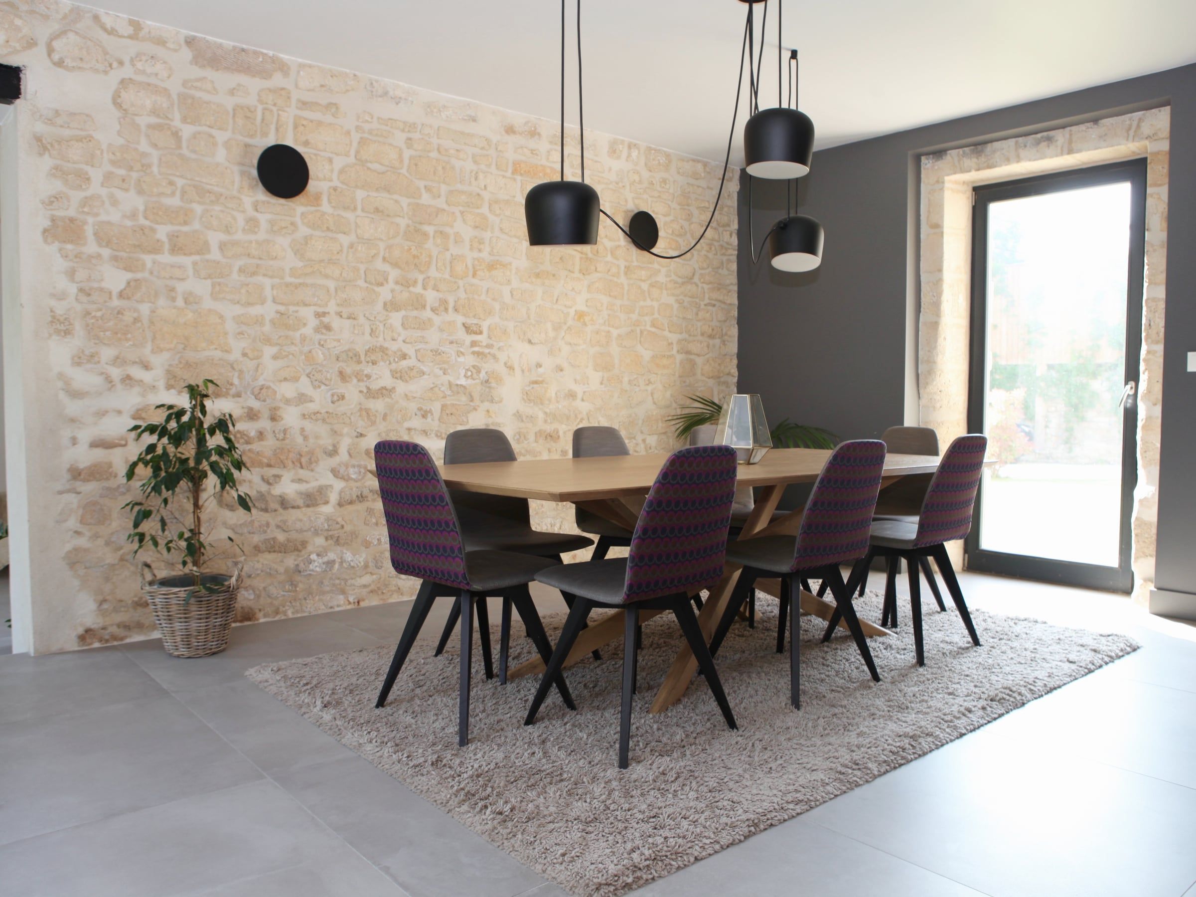 Salle à manger contemporaine installée devant un mur en pierre, avec table en bois, chaises tapissées et suspensions noires déportées.