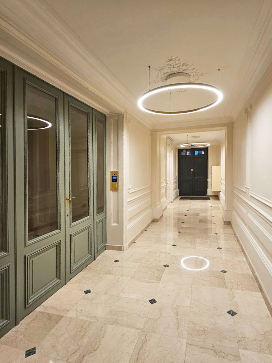 Hall d'immeuble haussmannien au sol en marbre à cabochons noirs, lustres circulaires LED suspendus, boiseries vert sauge et soubassements à moulures crème.