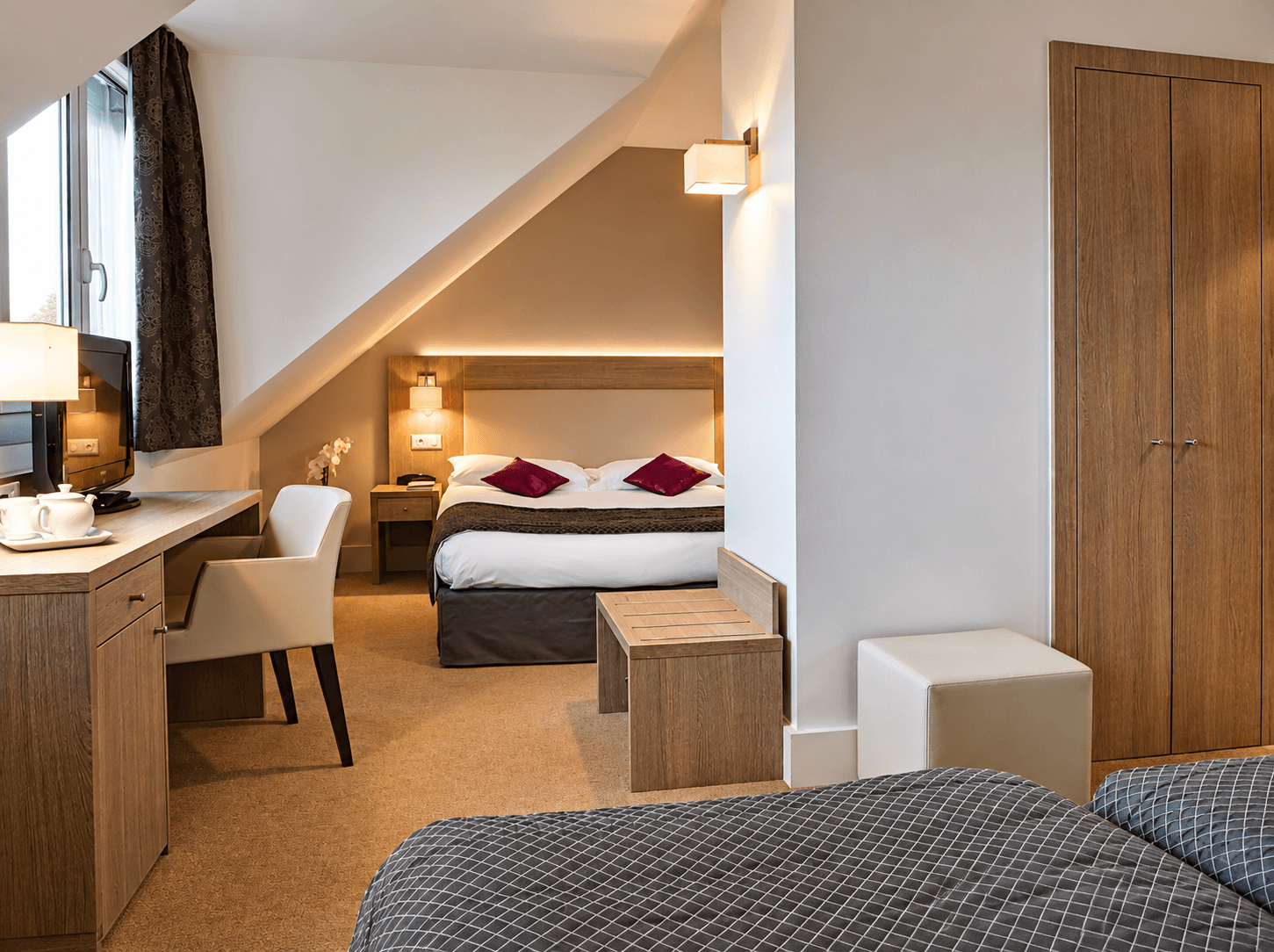 Chambre d'hôtel mansardée sous combles avec lit en bois clair, coussins prune, bureau en chêne et penderie à portes pleines.