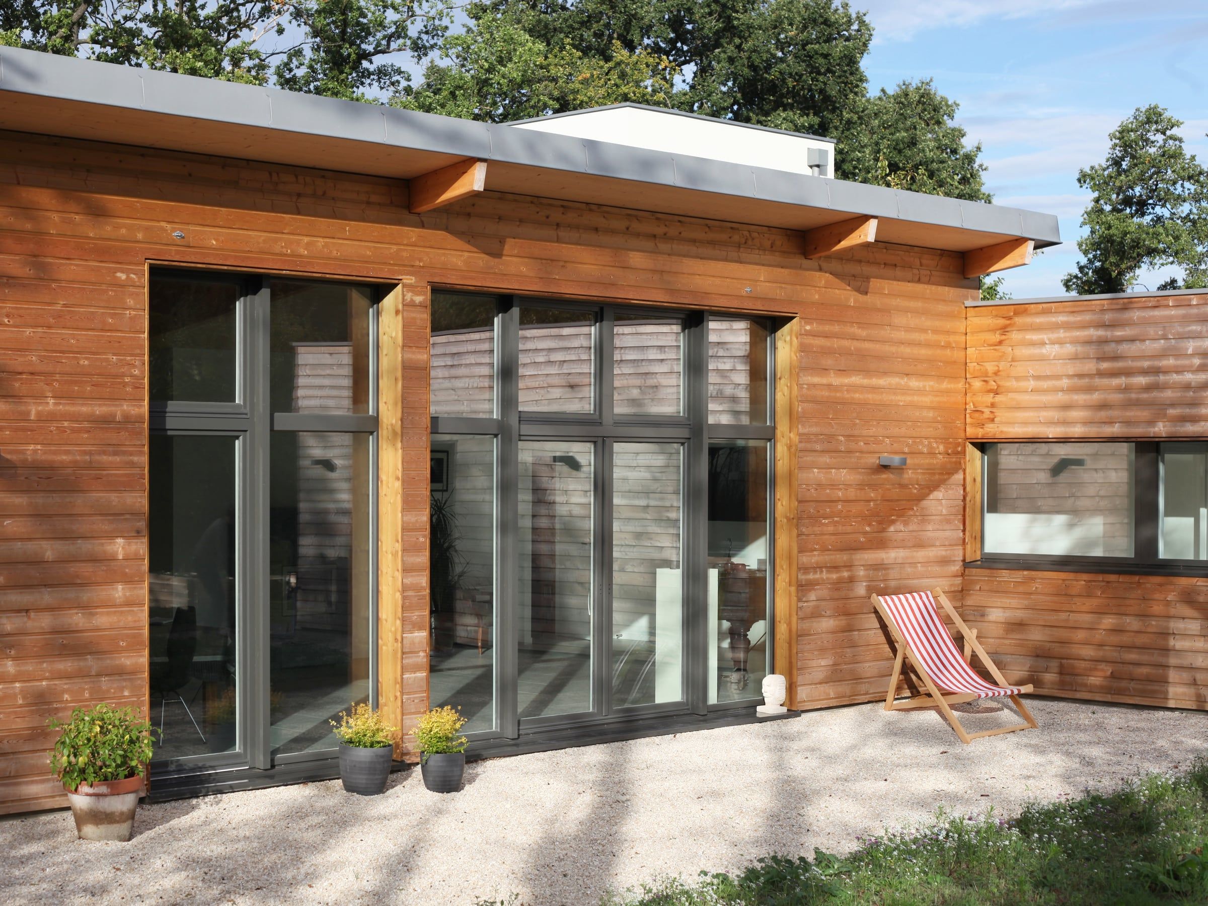 Aménagement de terrasse devant une façade en bardage bois, avec baies vitrées anthracite, débords de toiture et sol en gravier.