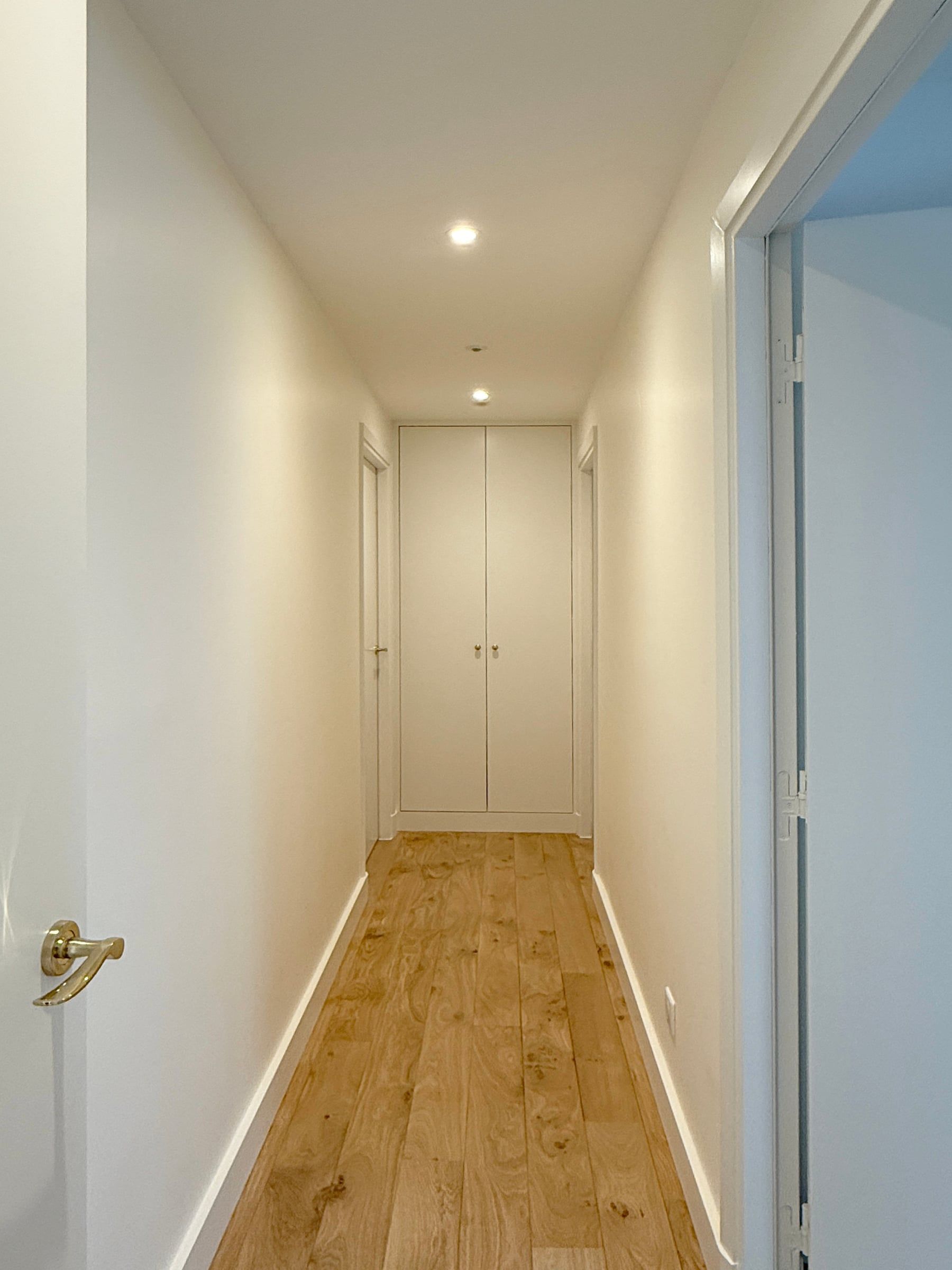 Couloir aux murs blancs et parquet en chêne clair, placard intégré toute hauteur et spots encastrés, agencement sur mesure rythmant la perspective.