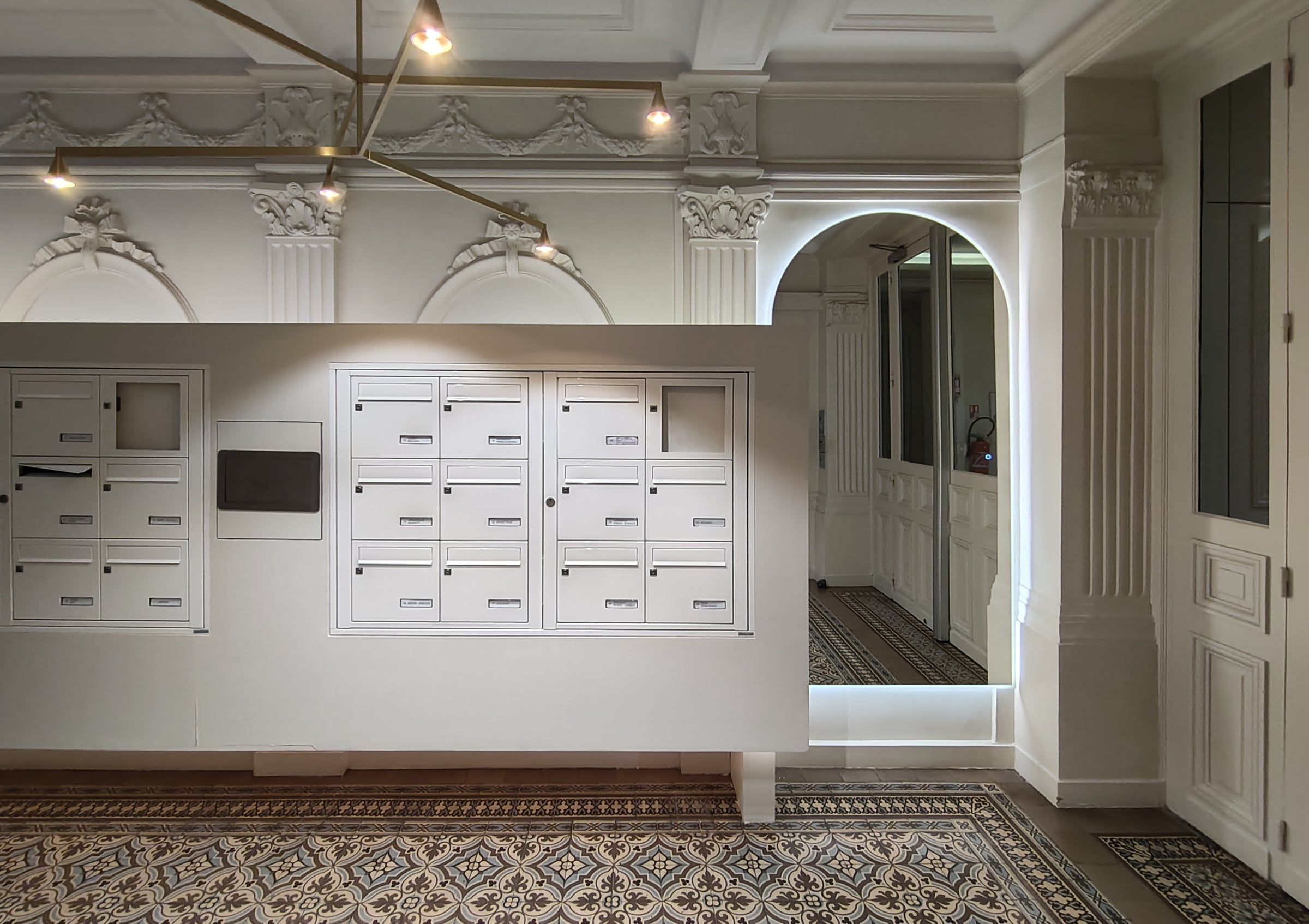 Hall haussmannien rénové par un architecte d'intérieur à Paris : boîtes aux lettres laquées blanches, carreaux de ciment, moulures, suspension en laiton.