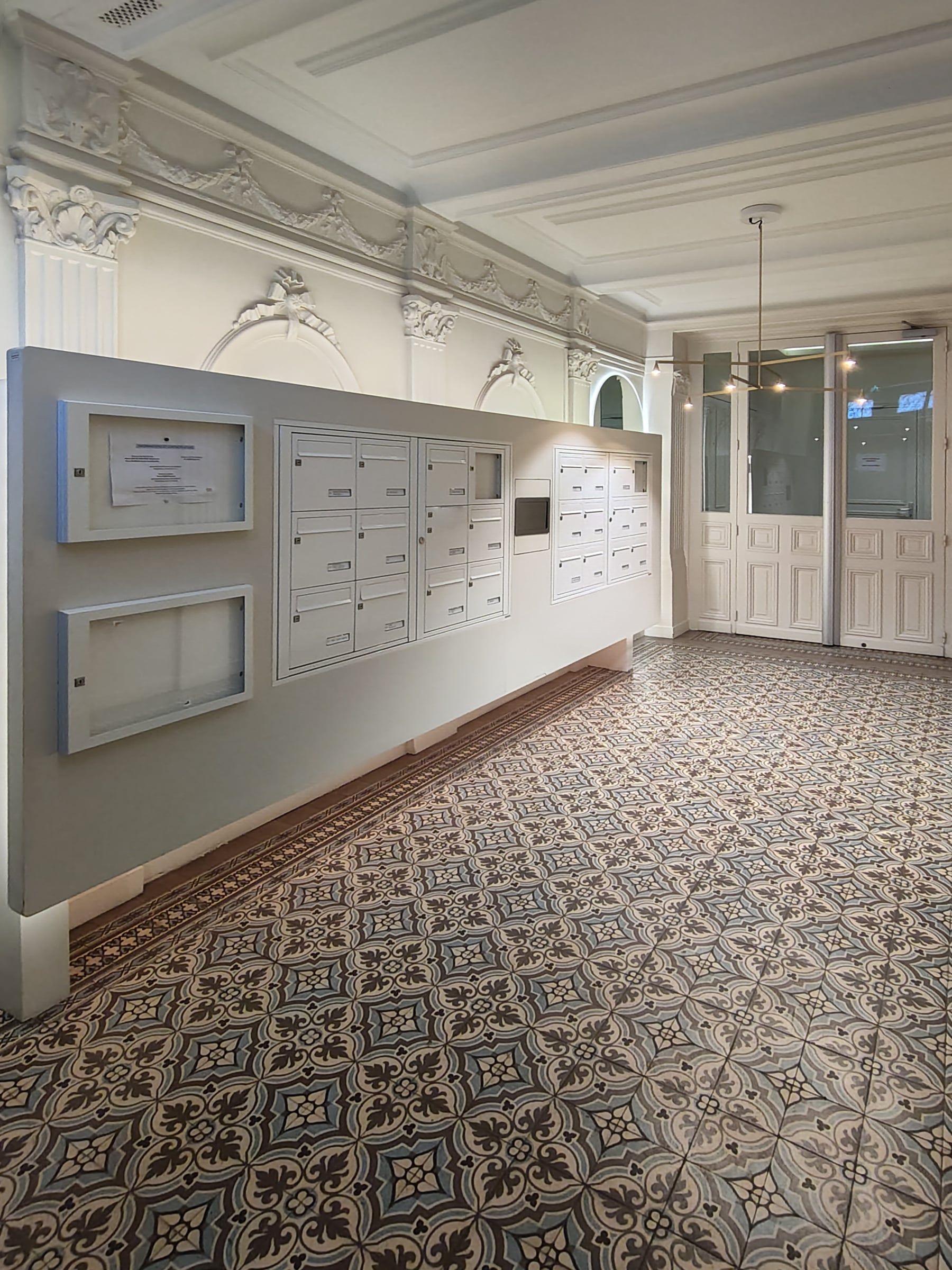 Hall aux moulures et stucs préservés, double rangée de boîtes aux lettres, sol en carreaux de ciment à motifs et suspension contemporaine.