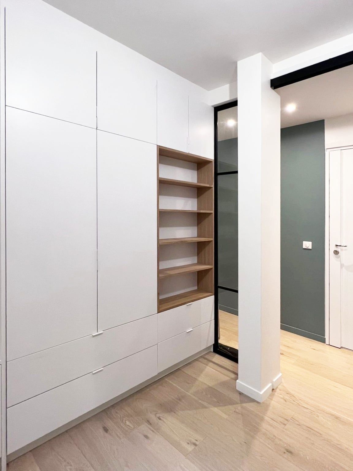 Dressing sur mesure aux façades blanches et niches en chêne, verrière noire ouvrant sur une entrée vert-de-gris au parquet clair, agencement sur mesure.