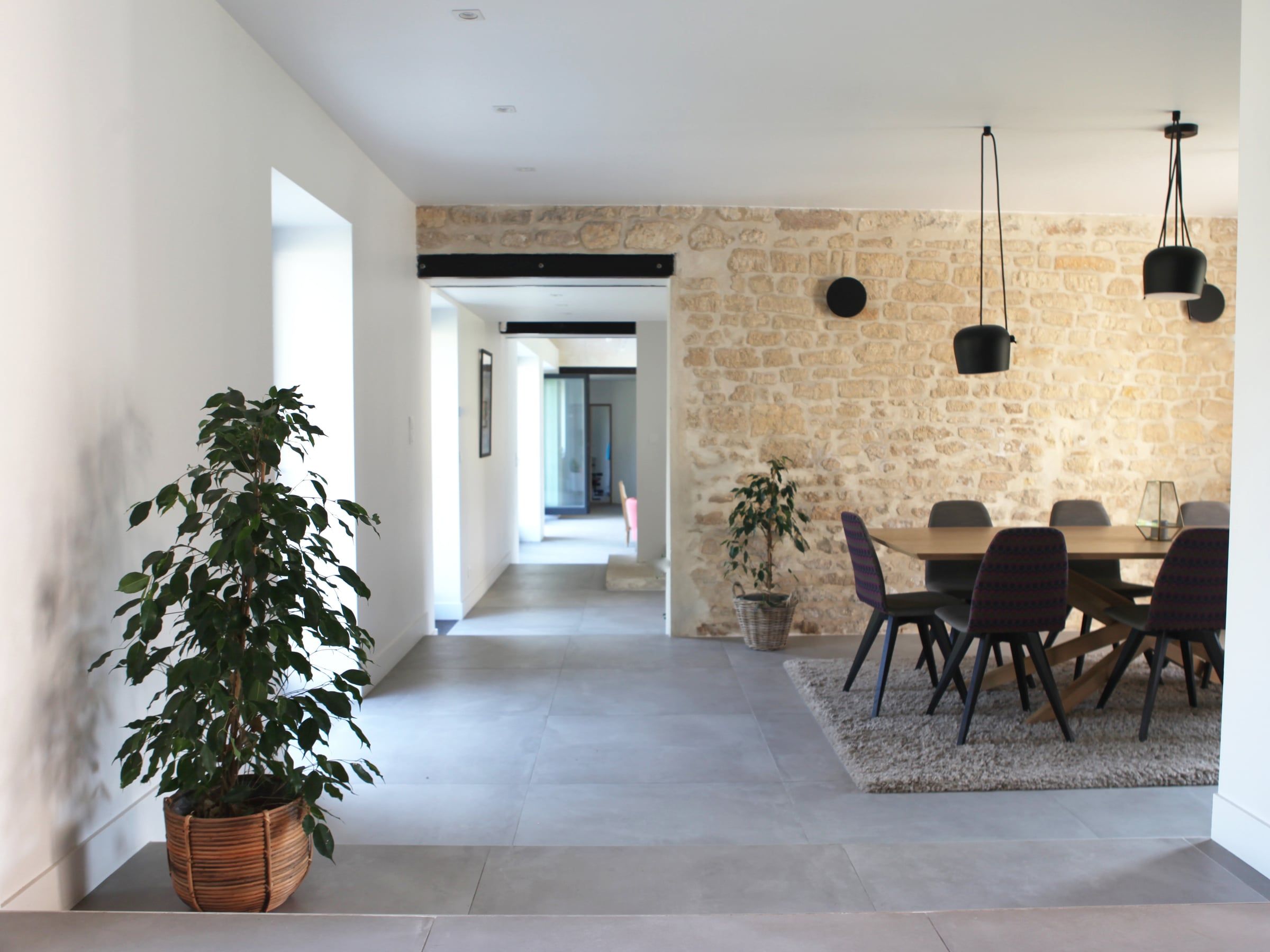 Rénovation de salle à manger ouverte sur la circulation, avec mur en pierre apparente, sol en grands carreaux gris et suspensions noires.