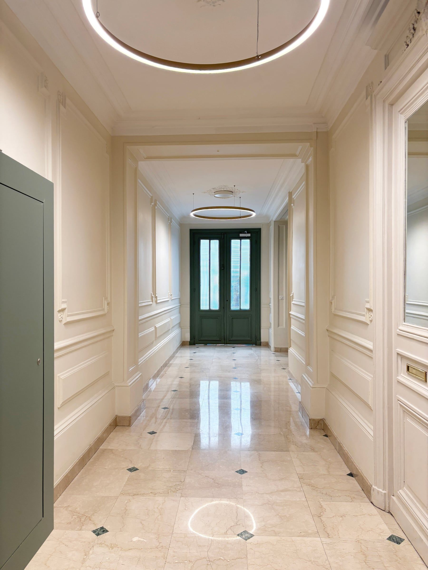 Couloir haussmannien au sol en marbre à cabochons, soubassements à moulures crème, lustres circulaires LED et double porte vitrée vert foncé en fond, hall d'immeuble.