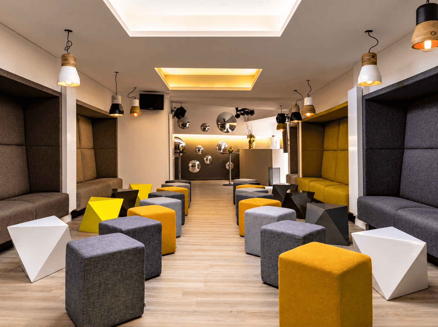 Salon de réception d'hôtel avec poufs géométriques jaunes et gris, banquettes à dossier haut, suspensions industrielles et parquet clair.