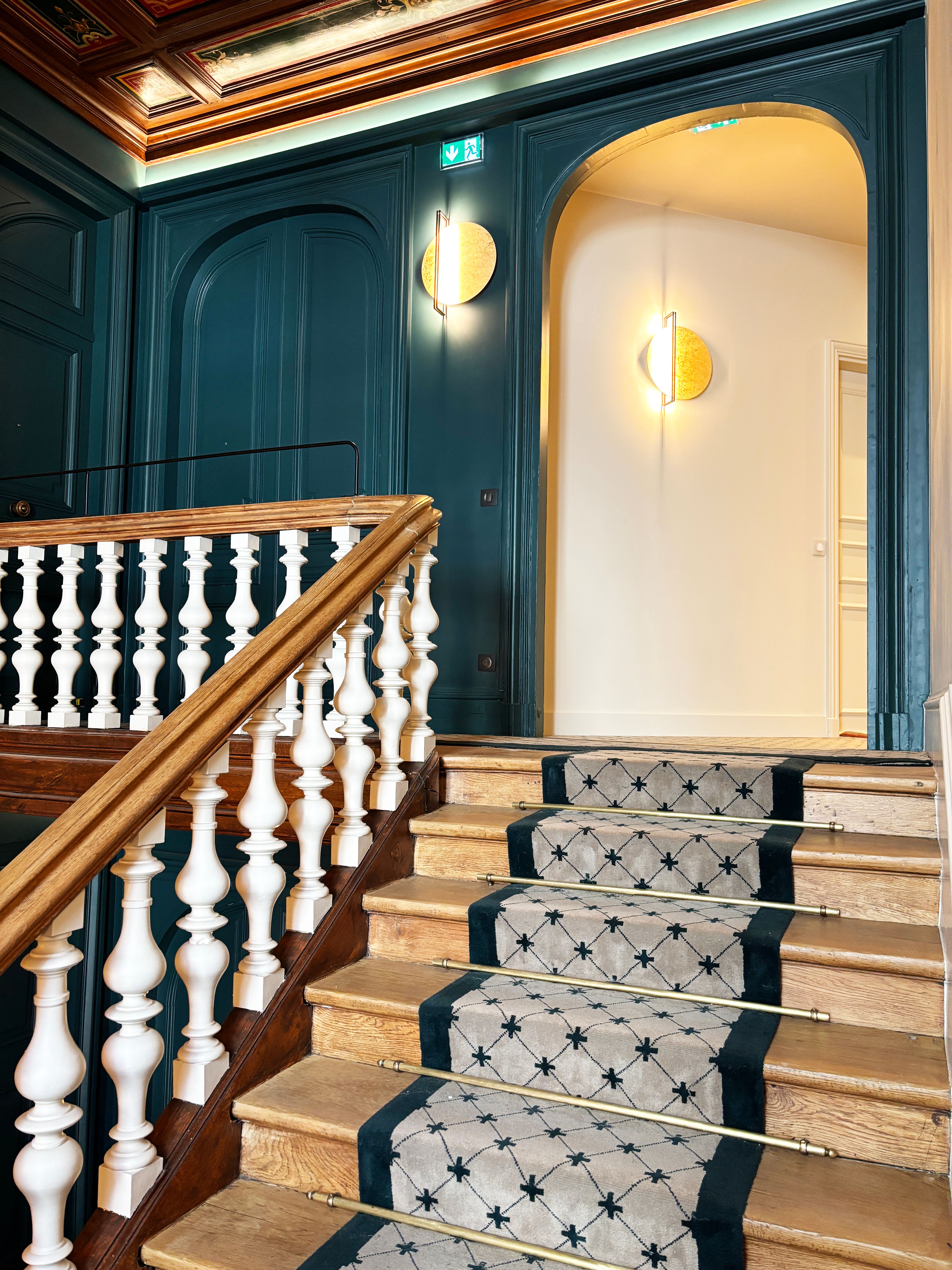 Montée d’escalier en bois avec tapis sur mesure et tringles métalliques dorées, encadrée par portes moulurées et appliques murales.