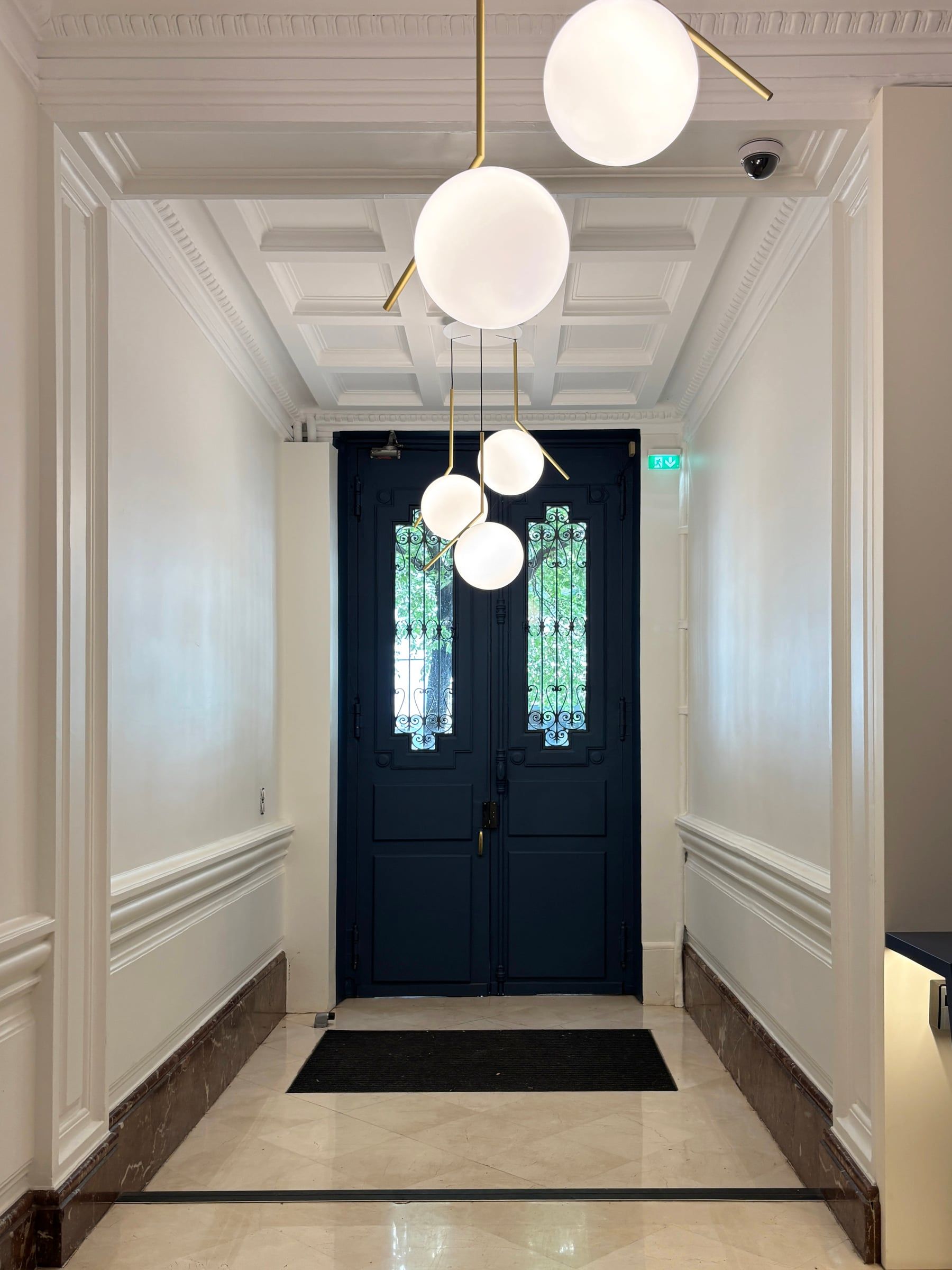 Entrée d’immeuble avec double porte bleu nuit vitrée, ferronneries décoratives, plafond à caissons et éclairage suspendu.