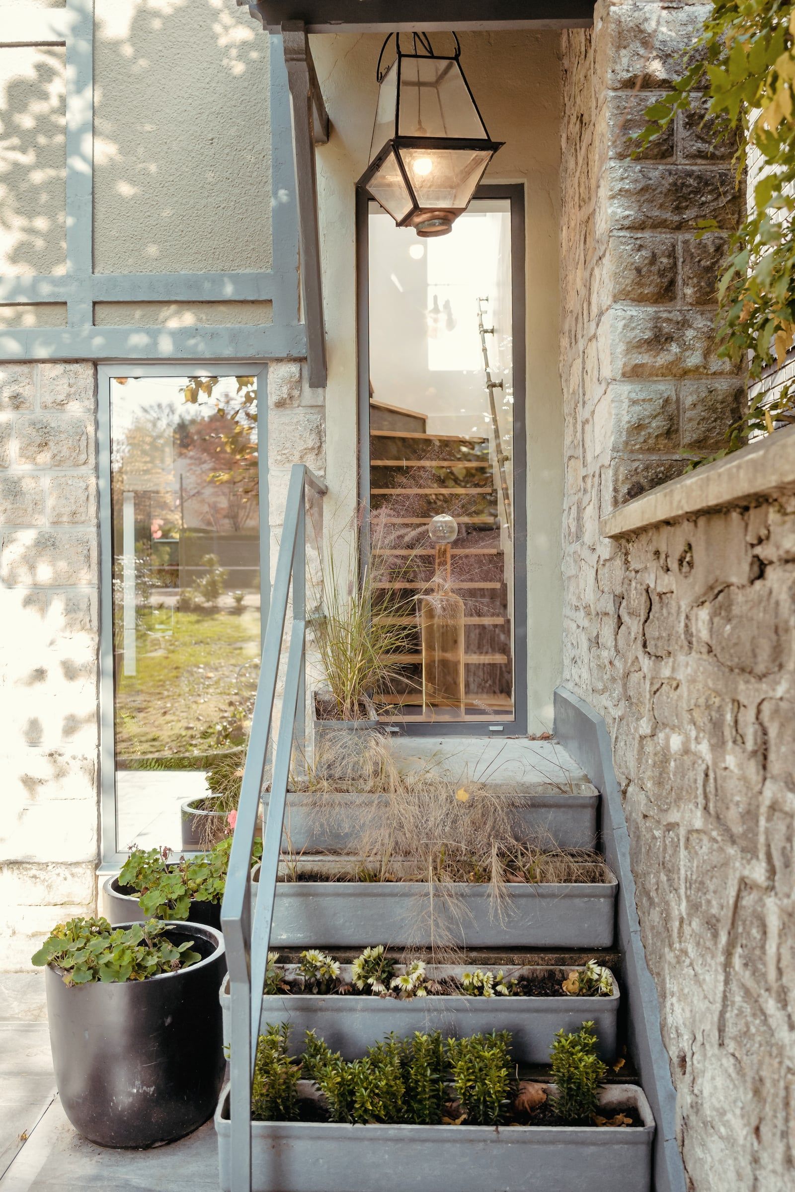 Escalier extérieur en pierre bordé de jardinières plantées, rampe métallique et lanterne suspendue contre un mur en moellons, rénovation de maison.