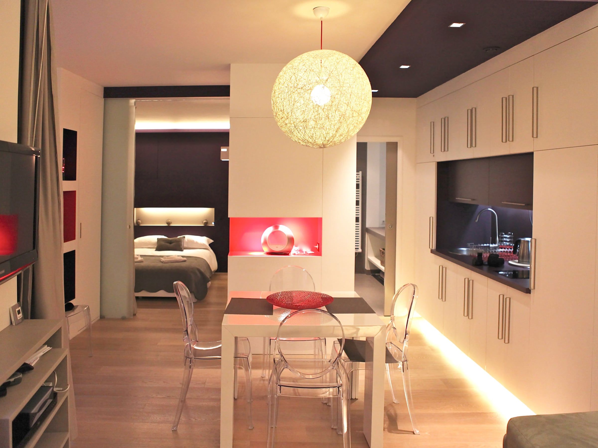 Studio contemporain ouvert reliant coin repas, chambre et cuisine, suspension boule et niche rouge rétroéclairée, rénové par un architecte d'intérieur.
