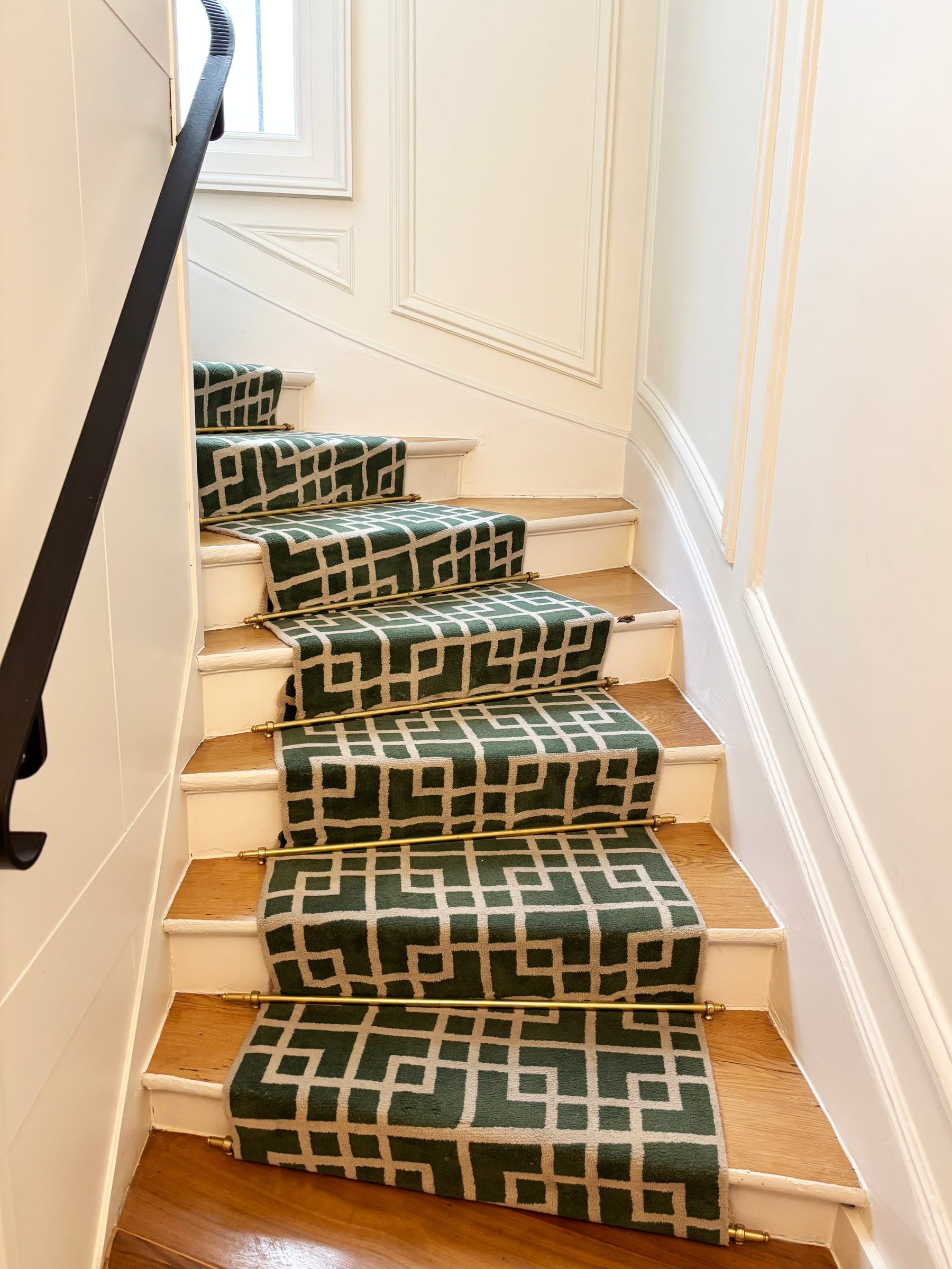 Cage d'escalier aux marches en chêne et chemin de tapis vert géométrique fixé par des barres en laiton, main courante en fer noir, rénovation de parties communes.