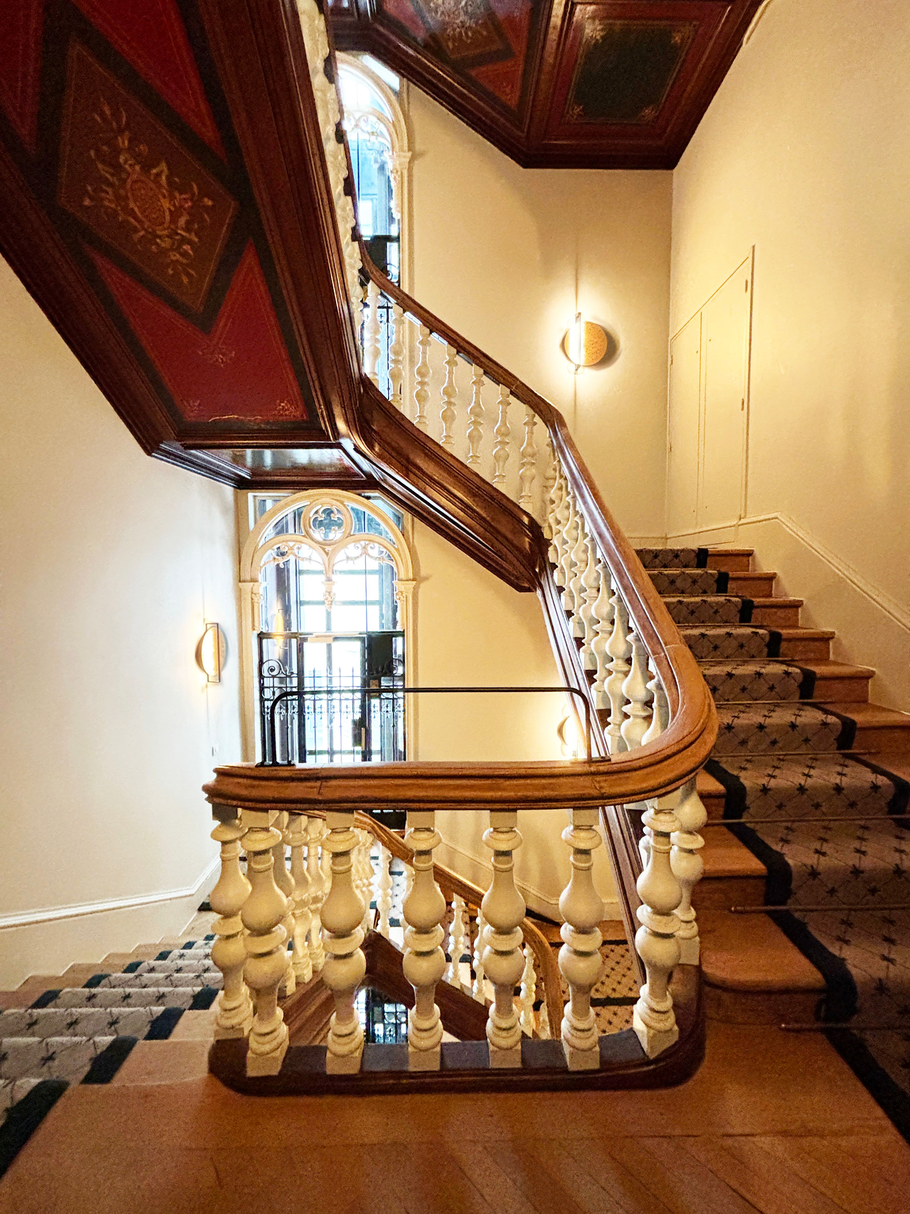Cage d’escalier éclairée par une grande baie cintrée, rampe en bois verni, balustres blancs et tapis posé sur marches en bois.
