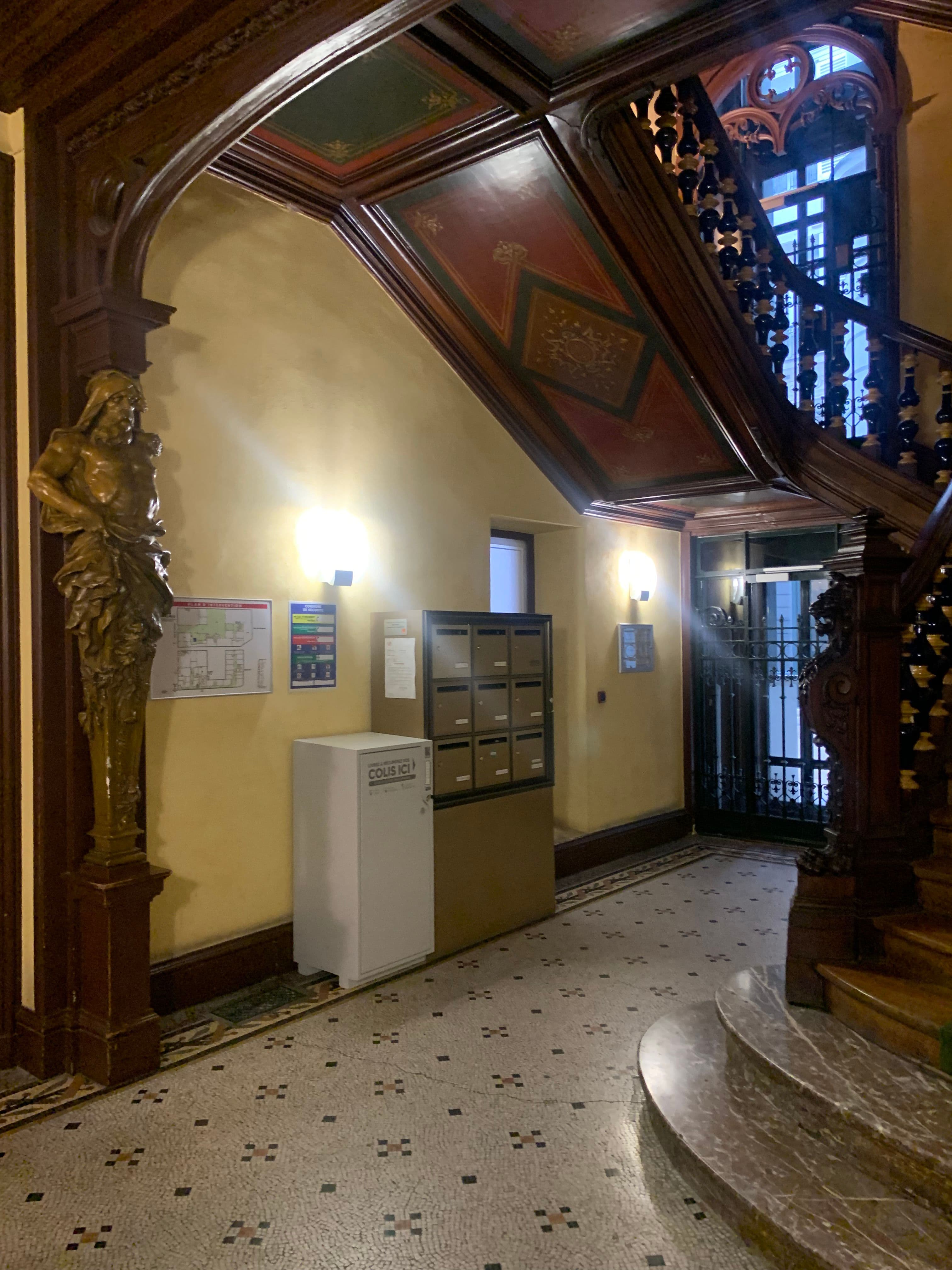 Avant travaux : hall d’immeuble aux murs jaunes, boîtes aux lettres apparentes, escalier en bois et décor peint en sous-face.