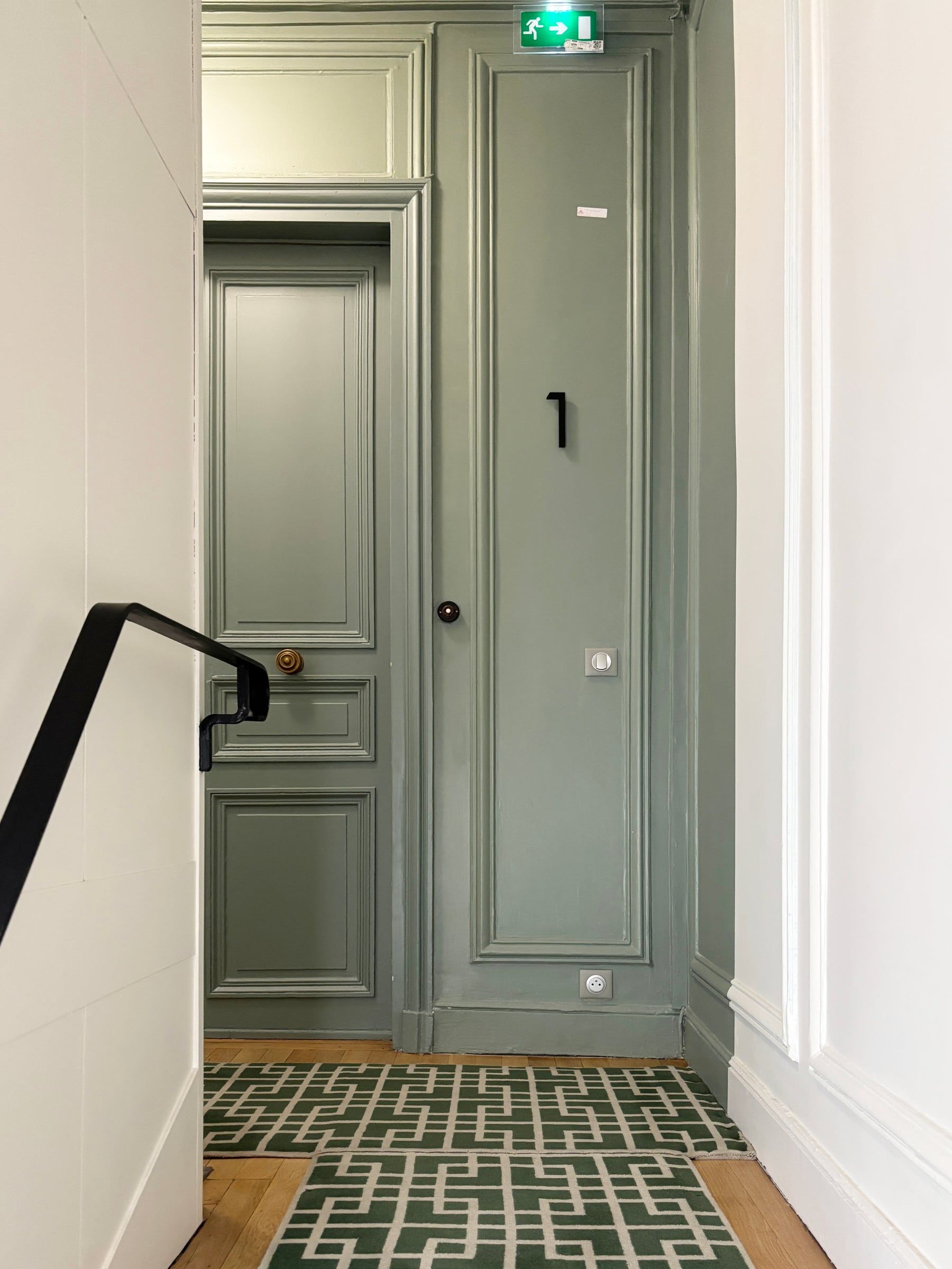 Palier haussmannien avec porte palière vert sauge à moulures et poignée en laiton, tapis vert géométrique sur parquet en chevrons et rampe noire, hall d'immeuble.