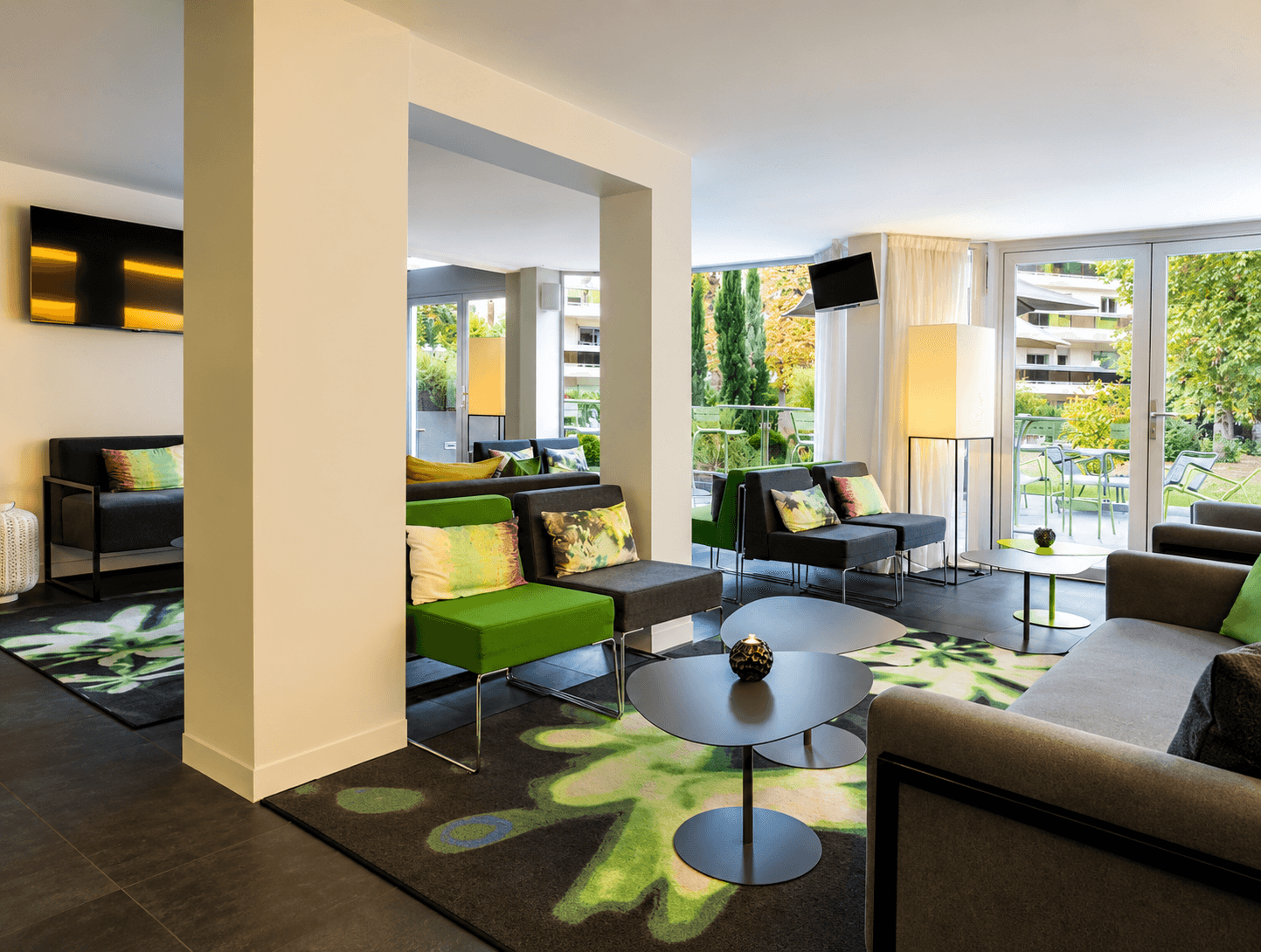 Salon lounge d'hôtel avec fauteuils gris et verts, guéridons ovales, tapis à motif floral et baies vitrées ouvrant sur le jardin.