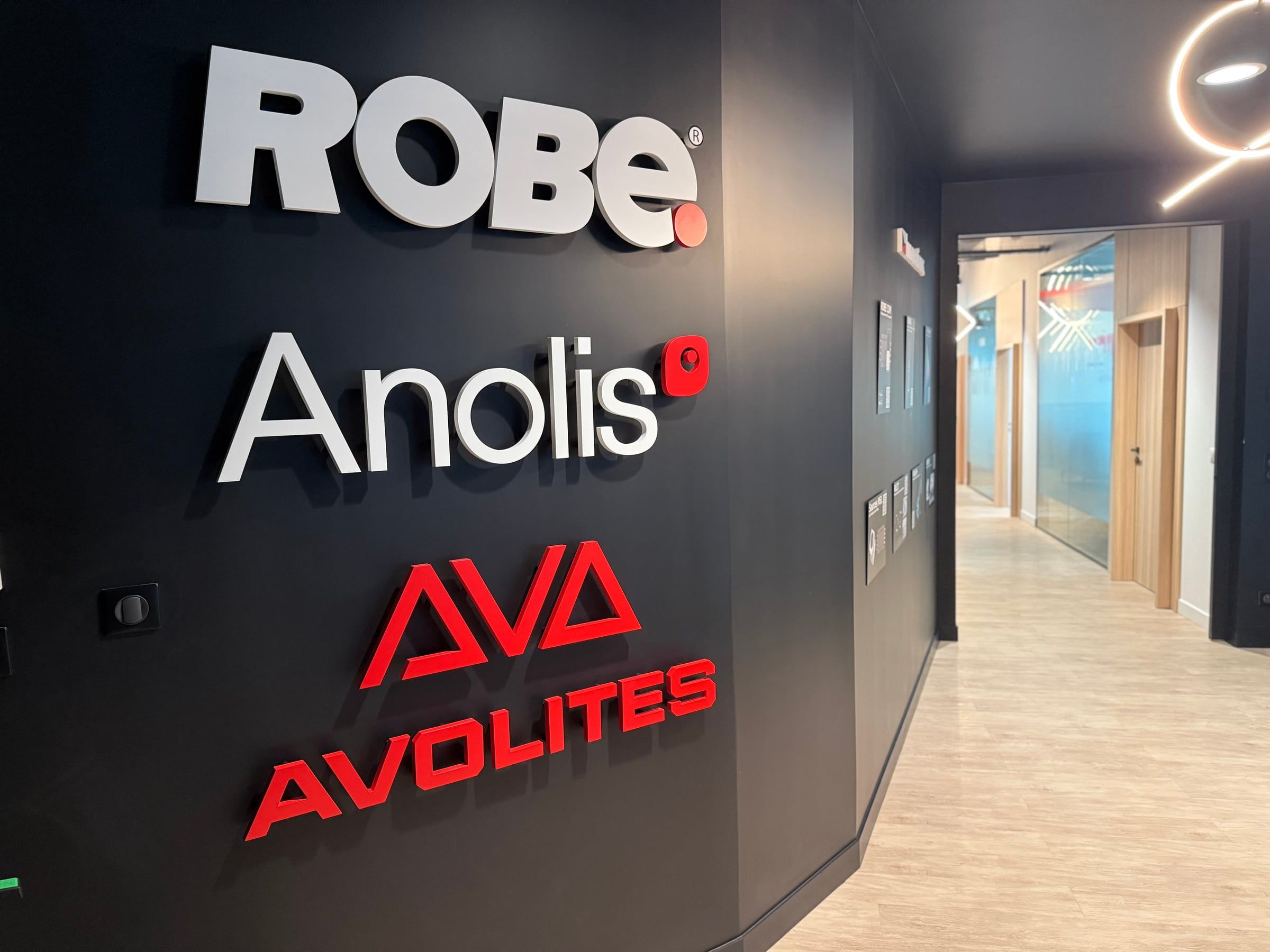 Entrée d'espace de travail, mur noir signalétique aux logos Robe, Anolis et Avolites en relief, ouvrant sur un couloir habillé de chêne clair.