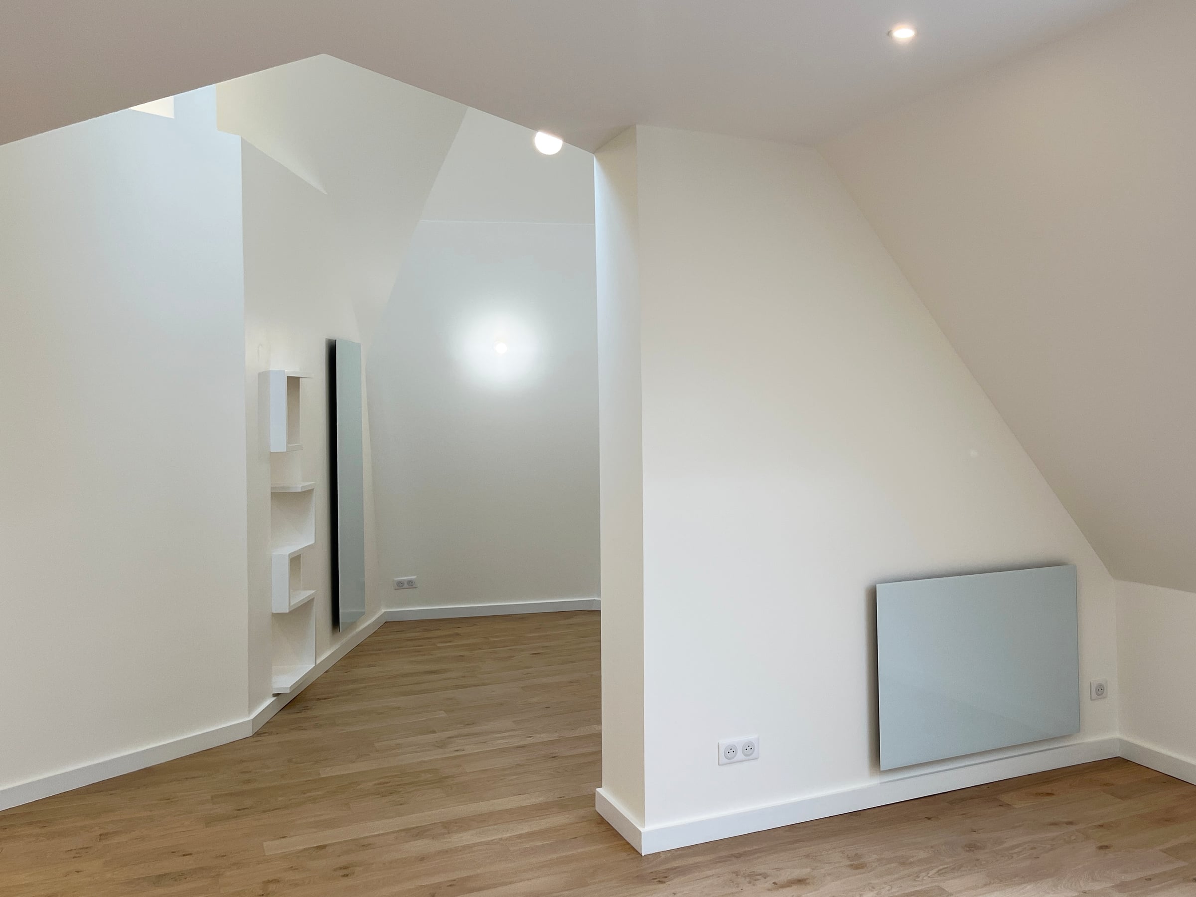 Espace de vie sous rampants, parquet en chêne clair, étagères blanches encastrées et radiateur design issus d'une rénovation d'appartement.