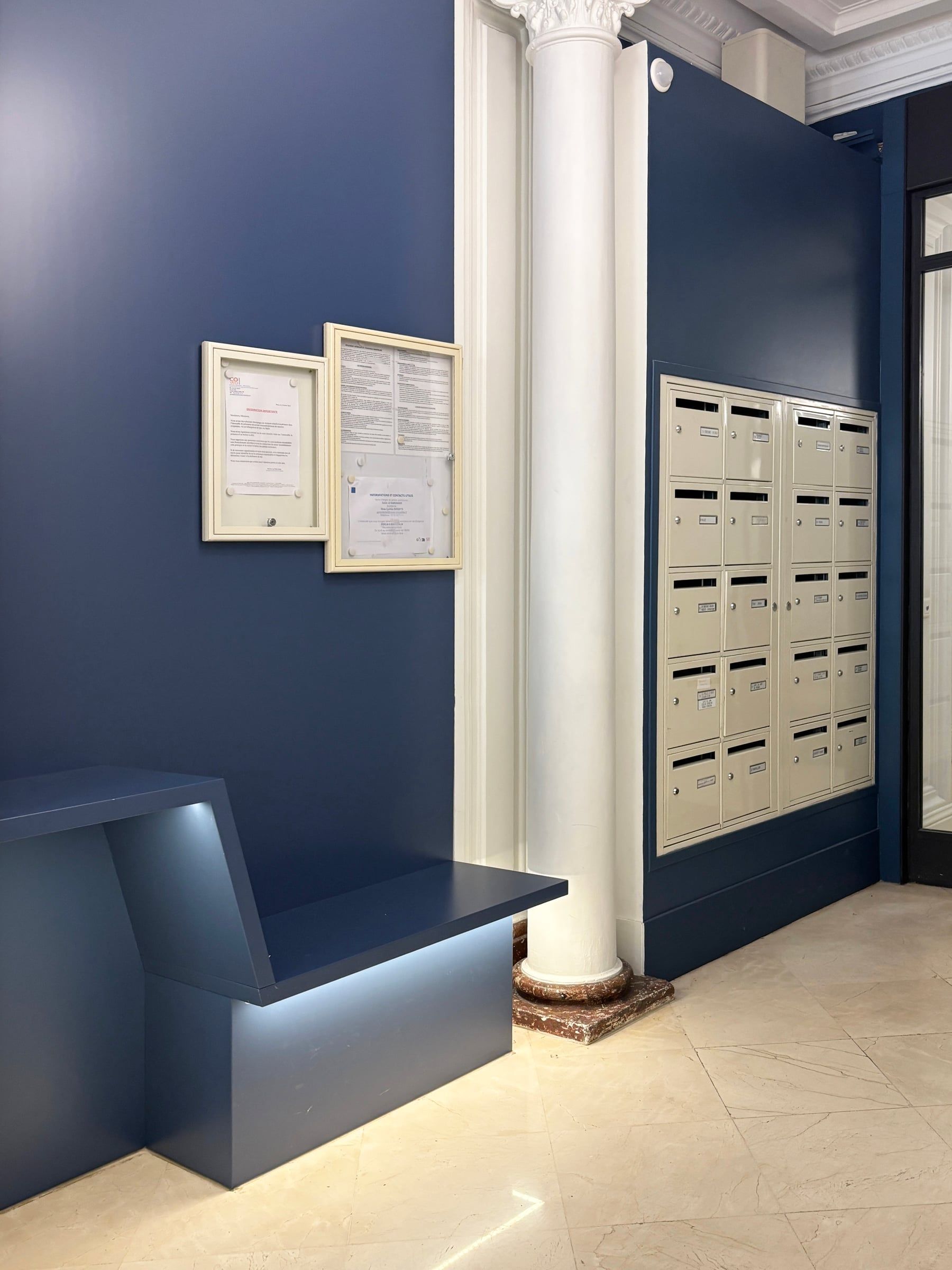 Agencement de hall d’immeuble intégrant boîtes aux lettres encastrées, panneau d’affichage et banc bleu éclairé en sous-face.