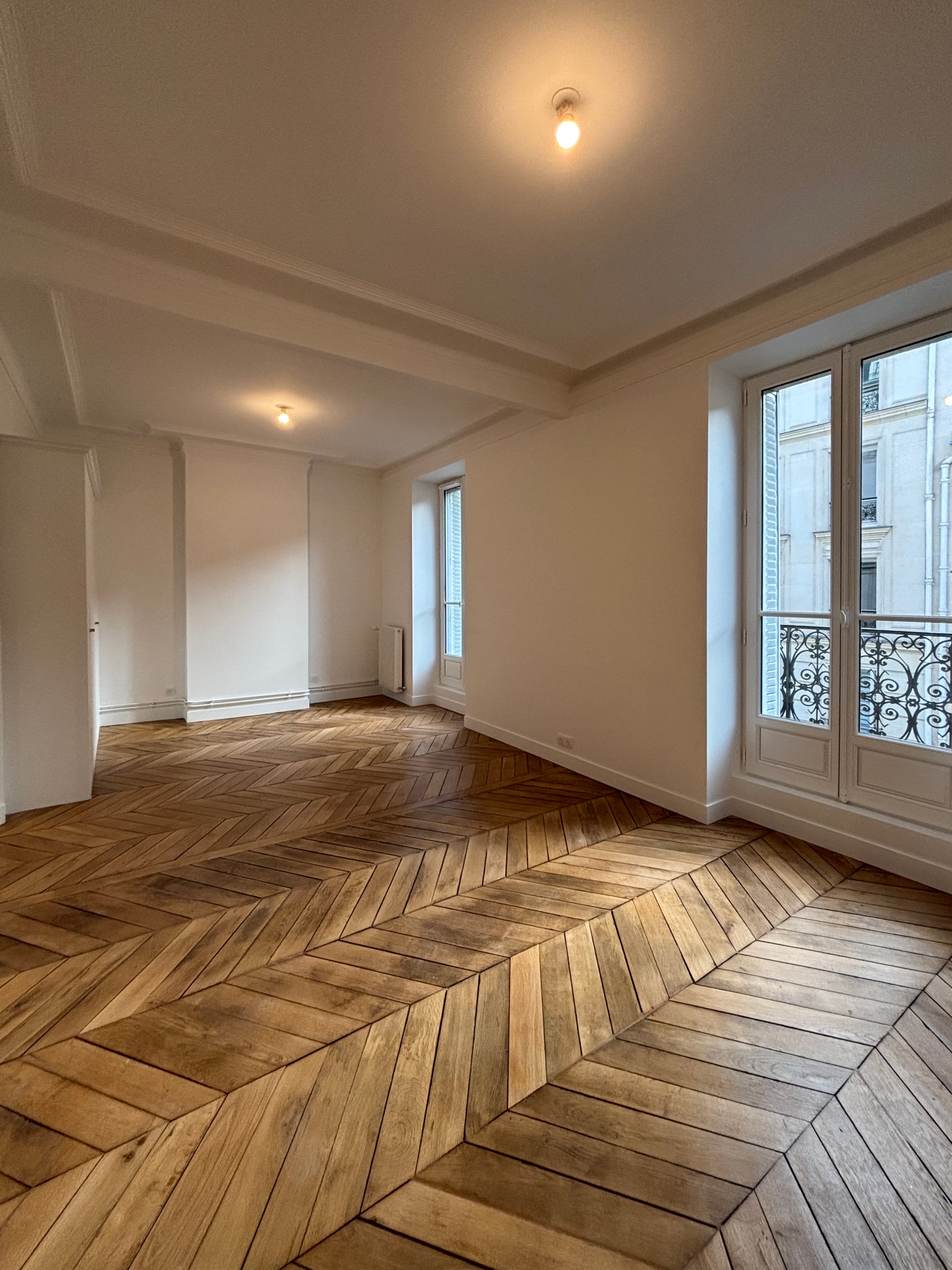 Séjour haussmannien aux poutres apparentes et parquet point de Hongrie, double porte-fenêtre à garde-corps en fer forgé sur la rue.