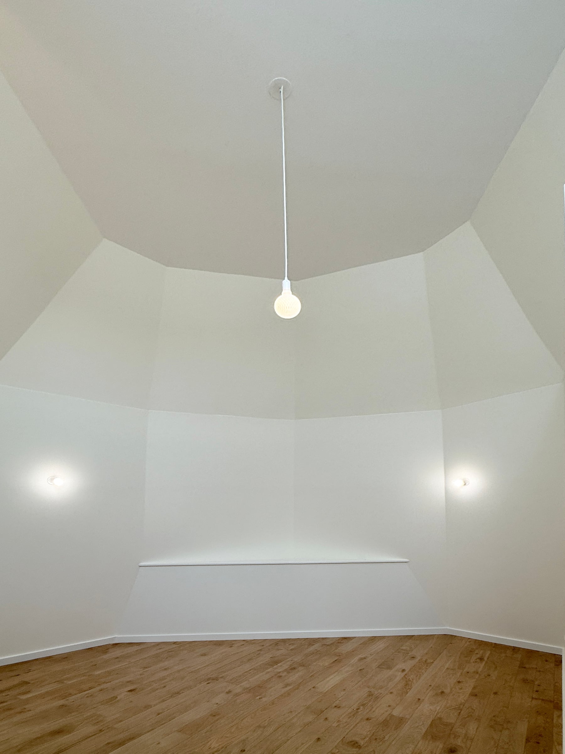 Chambre sous comble à plafond polygonal mansardé, parquet en chêne, suspension centrale et appliques murales sur murs blancs après rénovation.