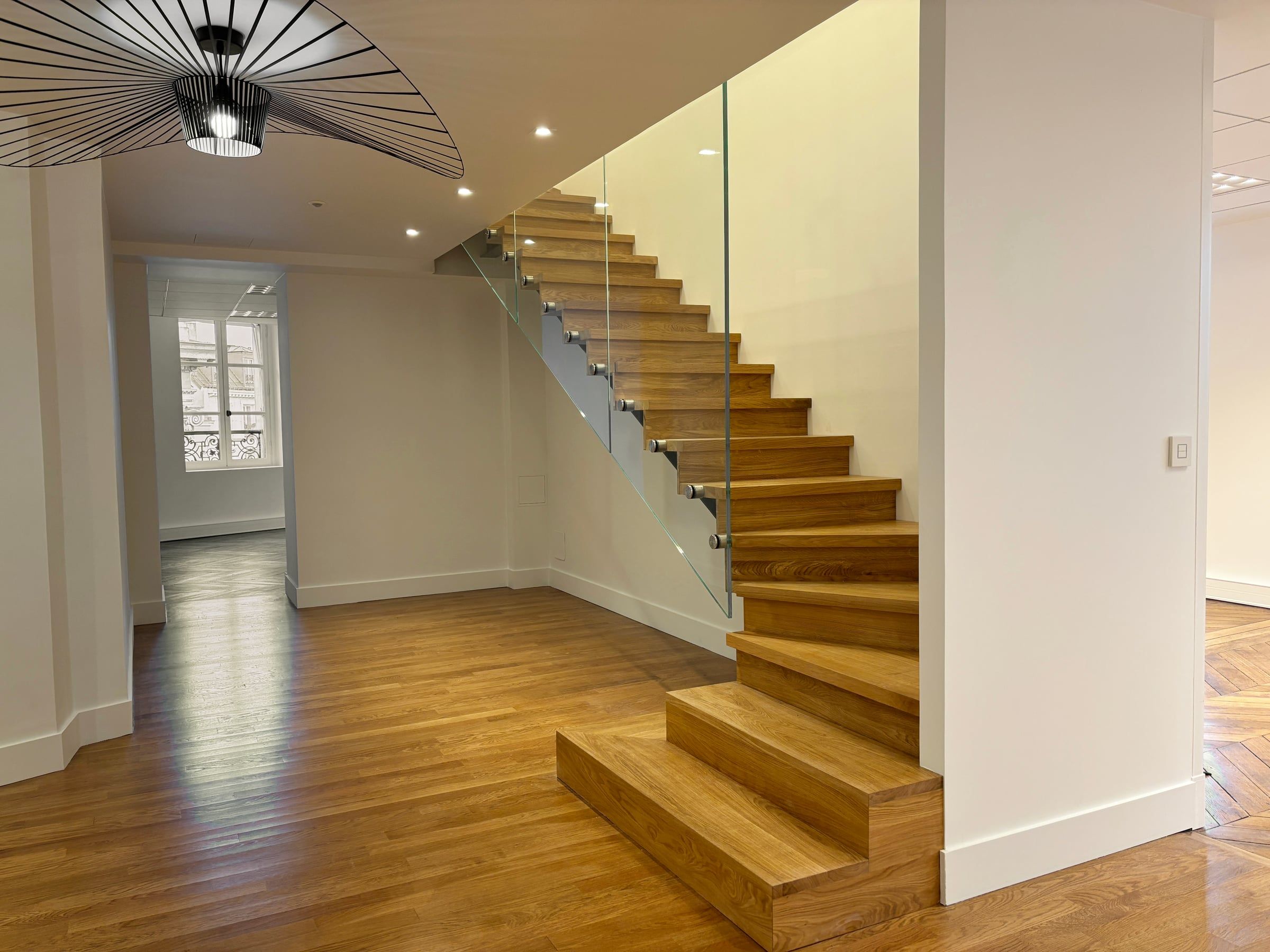 Hall de bureaux contemporain avec escalier suspendu en chêne, garde-corps en verre et suspension Vertigo noire, aménagé par un architecte d'intérieur tertiaire