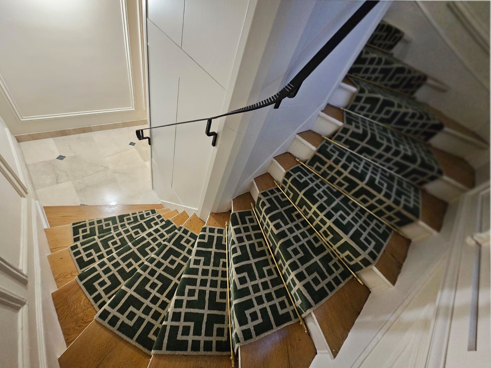Cage d'escalier en bois aux marches habillées d'un tapis géométrique vert et blanc, main courante en fer et sol en marbre, rénovée par un architecte d'intérieur à Paris.