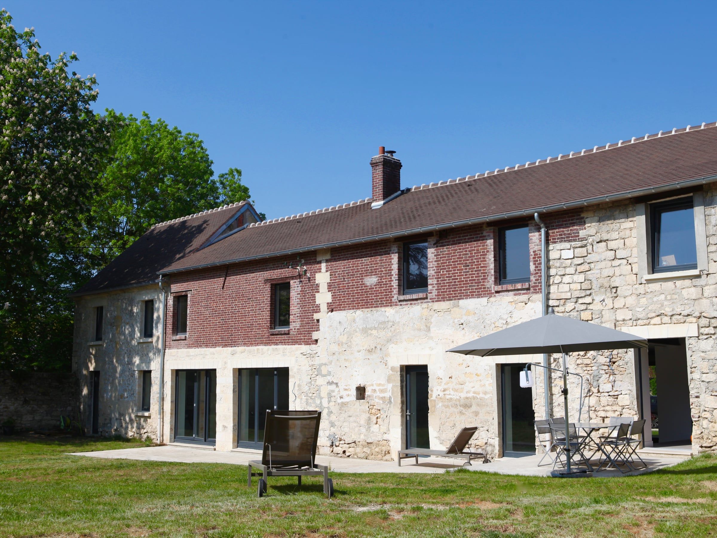 Maison ancienne rénovée par un architecte d’intérieur, associant façade en pierre et brique rouge, menuiseries sombres et terrasse sur jardin.