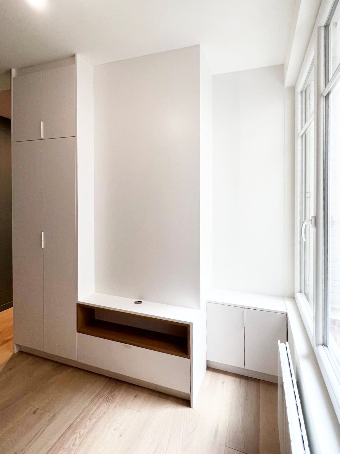 Meuble TV sur mesure en panneaux clairs avec niche en chêne intégrée, parquet en chêne et grande fenêtre à radiateur en fonte, réalisé sur mesure.