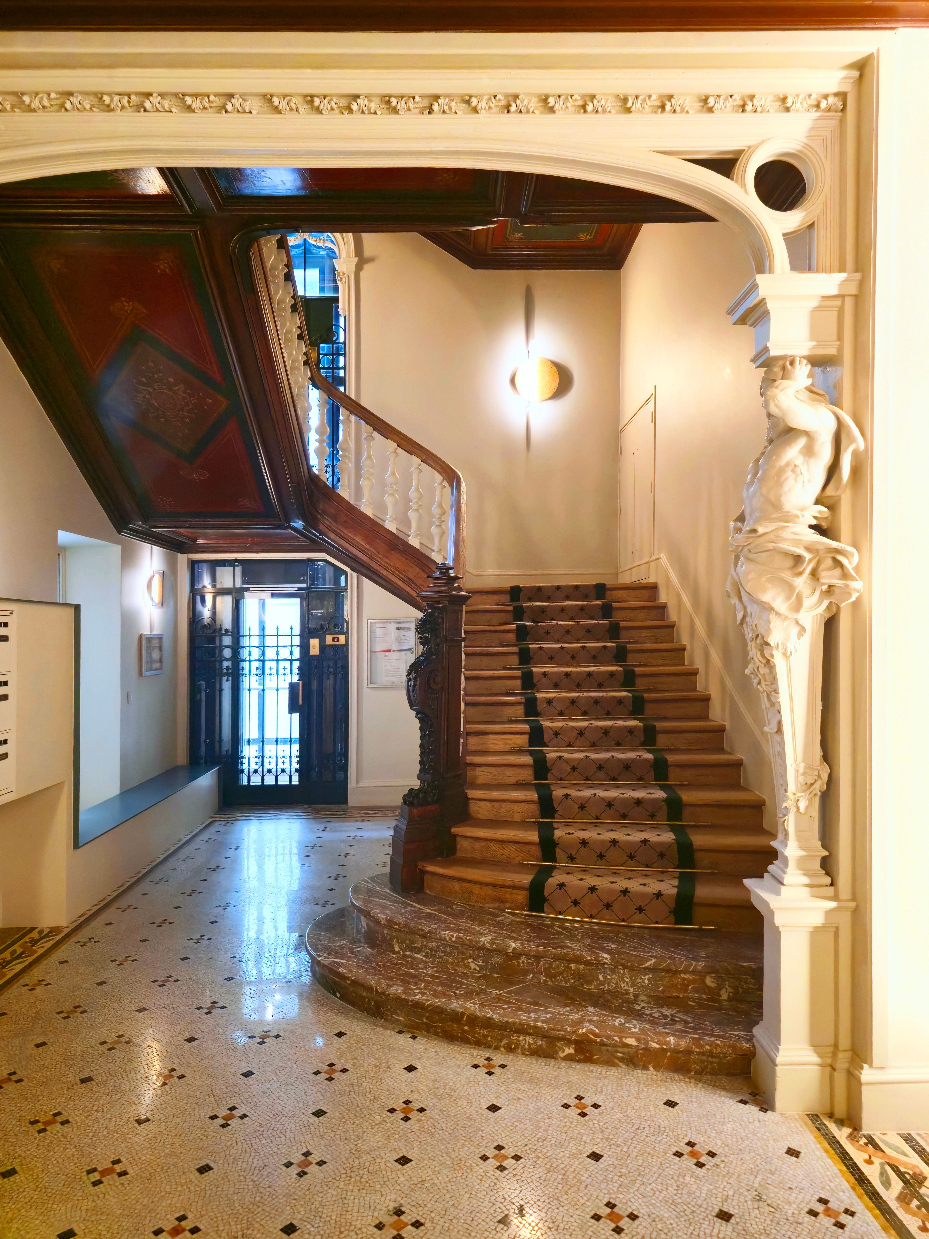 Hall d’entrée classé avec escalier en bois, départ en marbre, tapis central sur mesure et sculpture intégrée au pilastre mouluré.